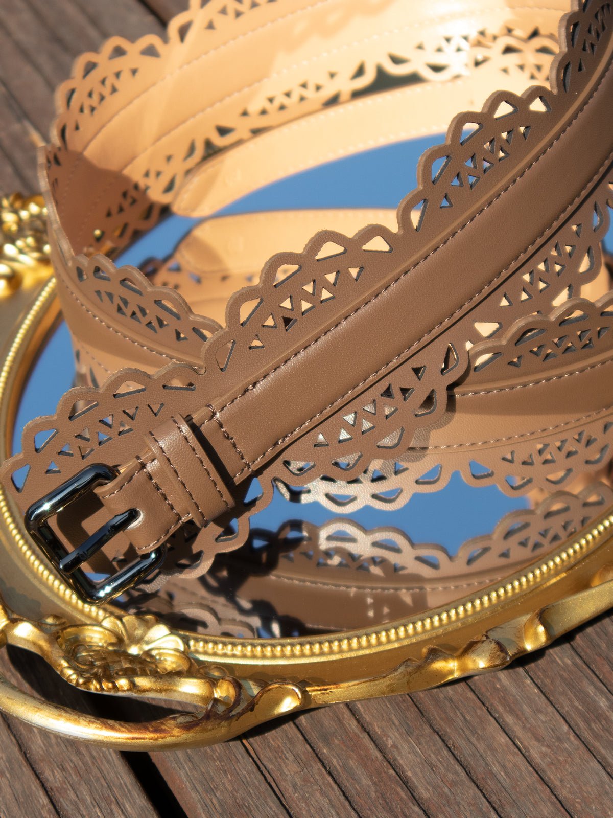 The Social Queen Bohemian Laser-Cut Leather Belt, featuring a metal buckle, is coiled atop an ornate gold-framed mirror on a wooden surface.