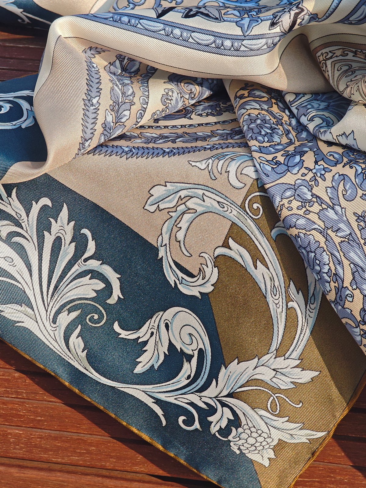 A close-up of the Social Queen Bohemian Print Silk Scarf, crafted from mulberry silk in blue, beige, and gold with ornate floral and leaf patterns, displayed on a wooden surface.