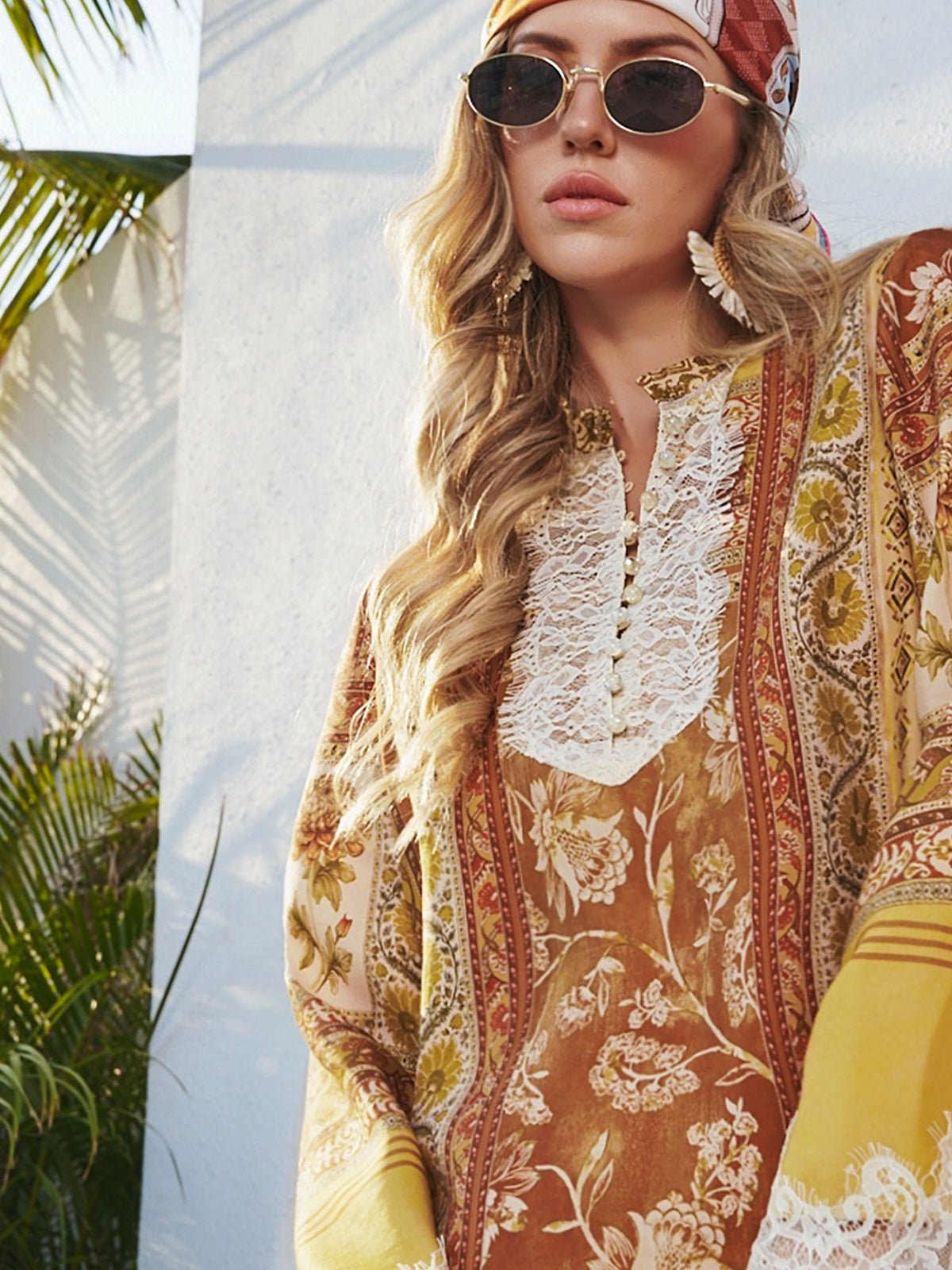 A woman in sunlight stands near a white wall and palm leaves, wearing Social Queen's Boho Lace Floral Print Dress along with sunglasses and a patterned headscarf.