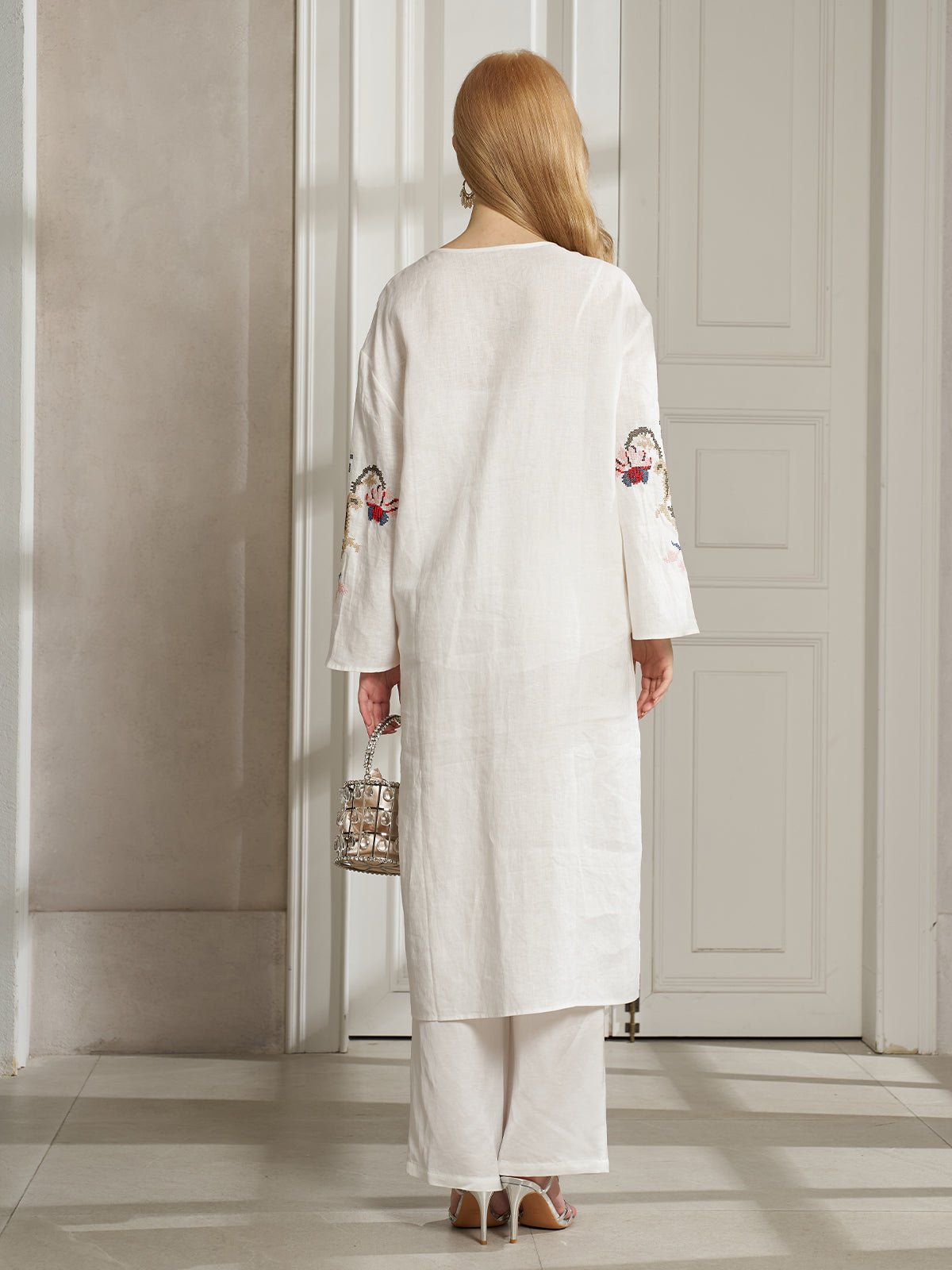 A woman with long blonde hair, wearing the Social Queen Botanical Embroidered Linen Top, stands indoors facing away in a resort-style white outfit, holding a small metallic handbag.