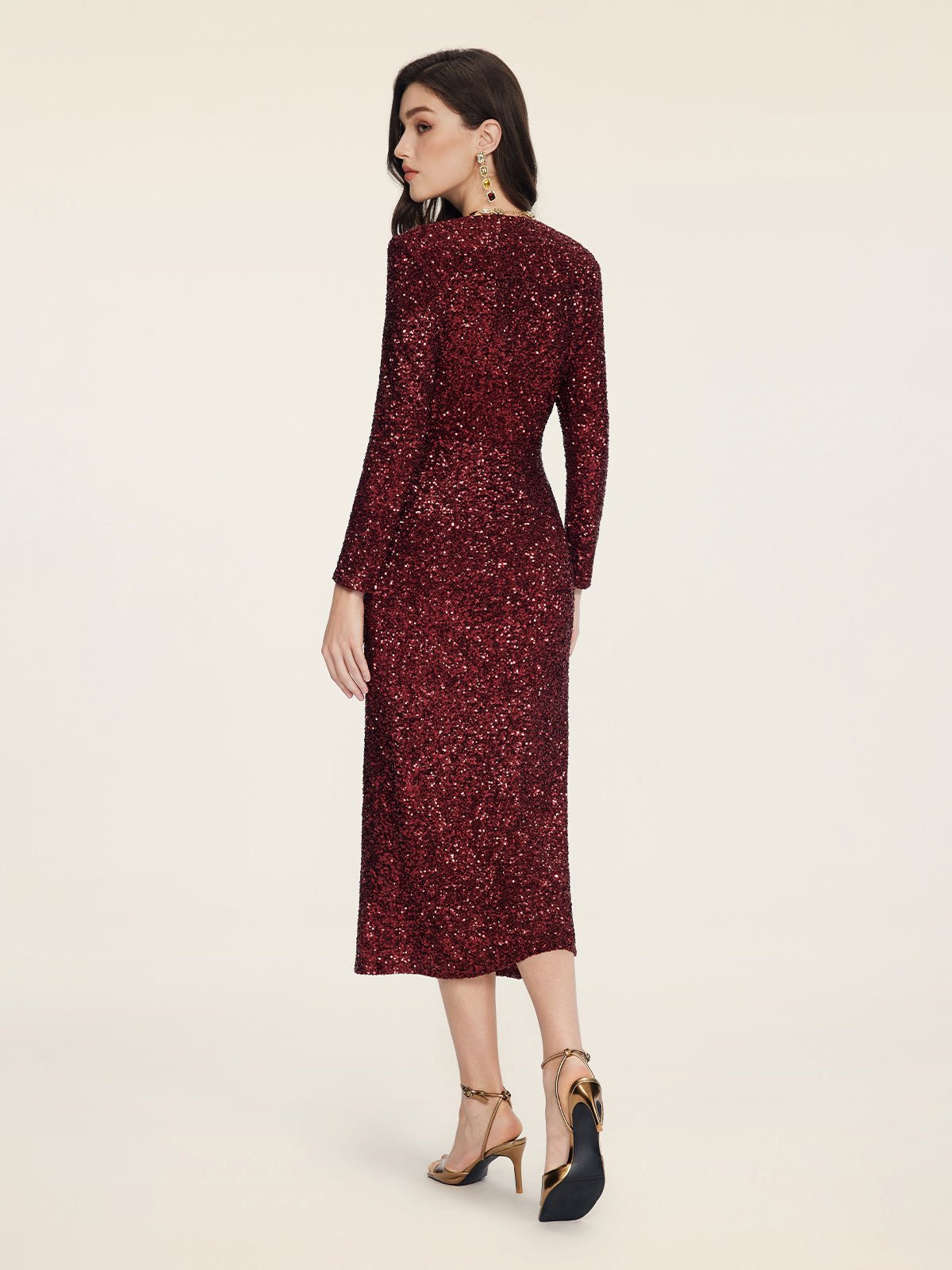 A woman in the Social Queen Burgundy Sequin V-Neck Slit Dress with gold heels stands against a plain light background, facing away from the camera.