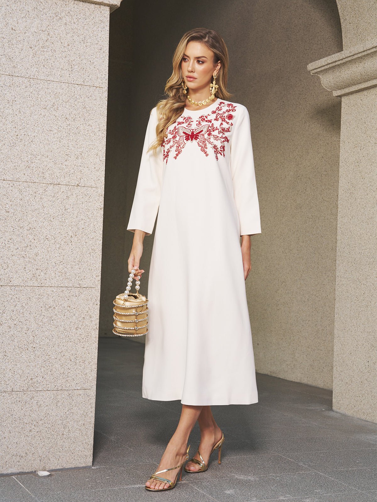 A woman stands near a stone wall wearing the Social Queen Butterfly Embroidered A-Line Dress, paired with gold sandals and a small gold handbag.