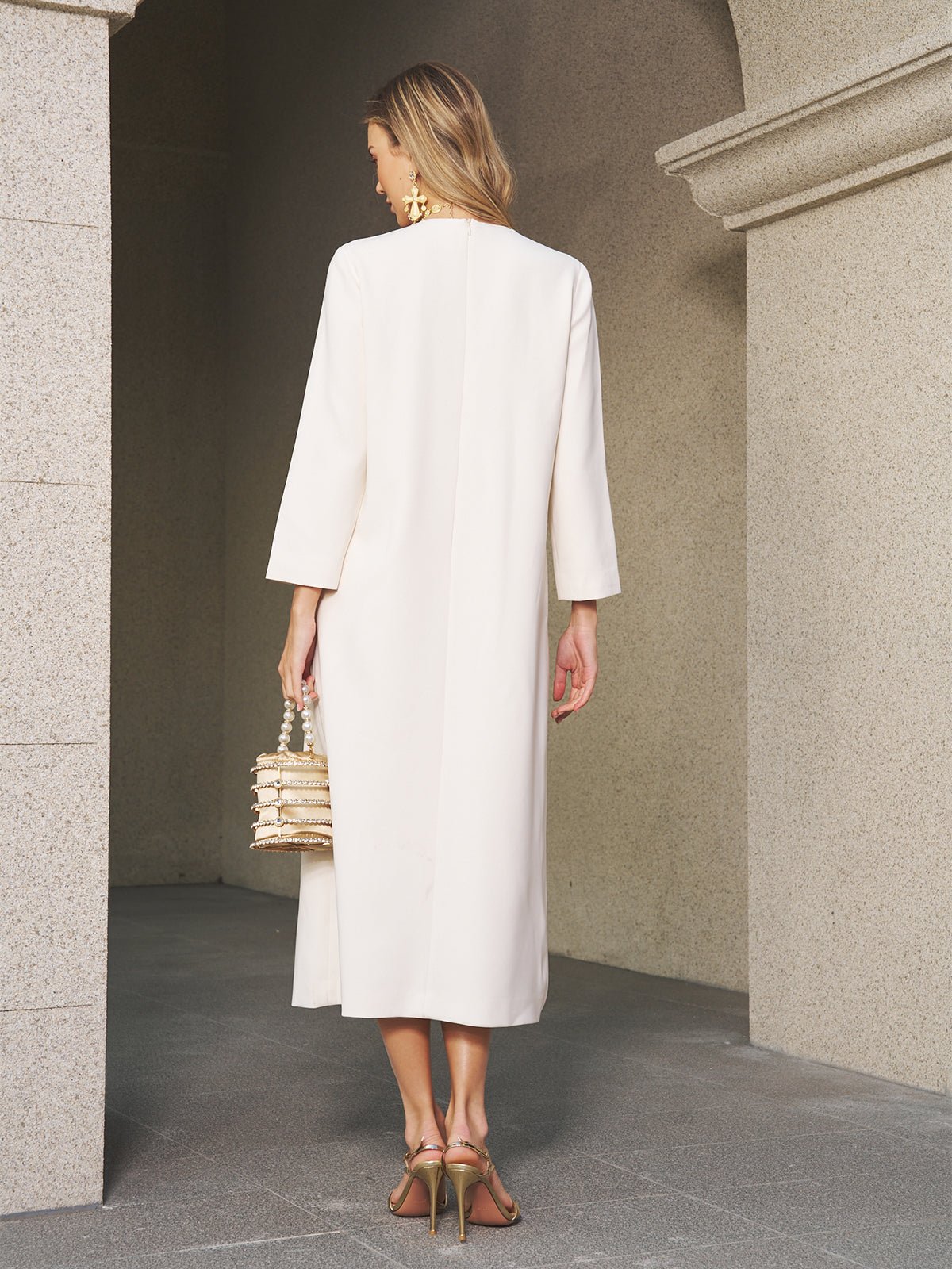 A woman wears the Social Queen Butterfly Embroidered A-Line Dress under a long cream coat, holding a textured handbag and sporting heeled sandals, her dress’s delicate floral and butterfly details visible in the architectural setting.