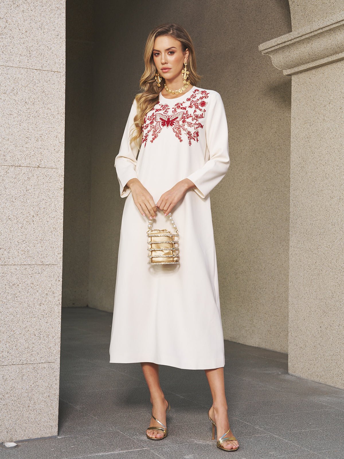 A woman in the Social Queen Butterfly Embroidered A-Line Dress stands by a stone wall, holding a gold handbag and wearing gold sandals with statement earrings.