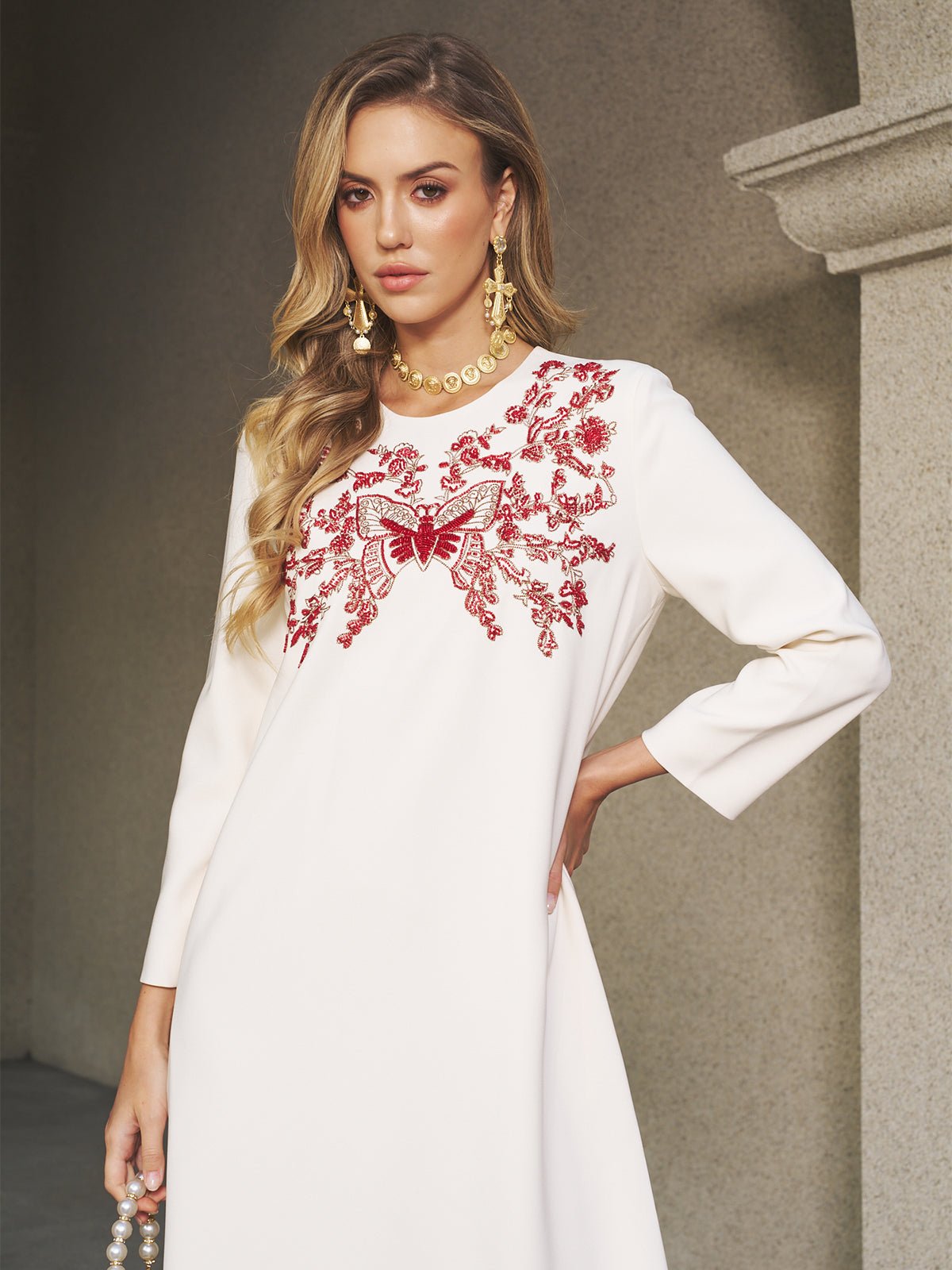A woman in the Social Queen Butterfly Embroidered A-Line Dress stands beside a stone wall, wearing gold jewelry and holding a beaded accessory.