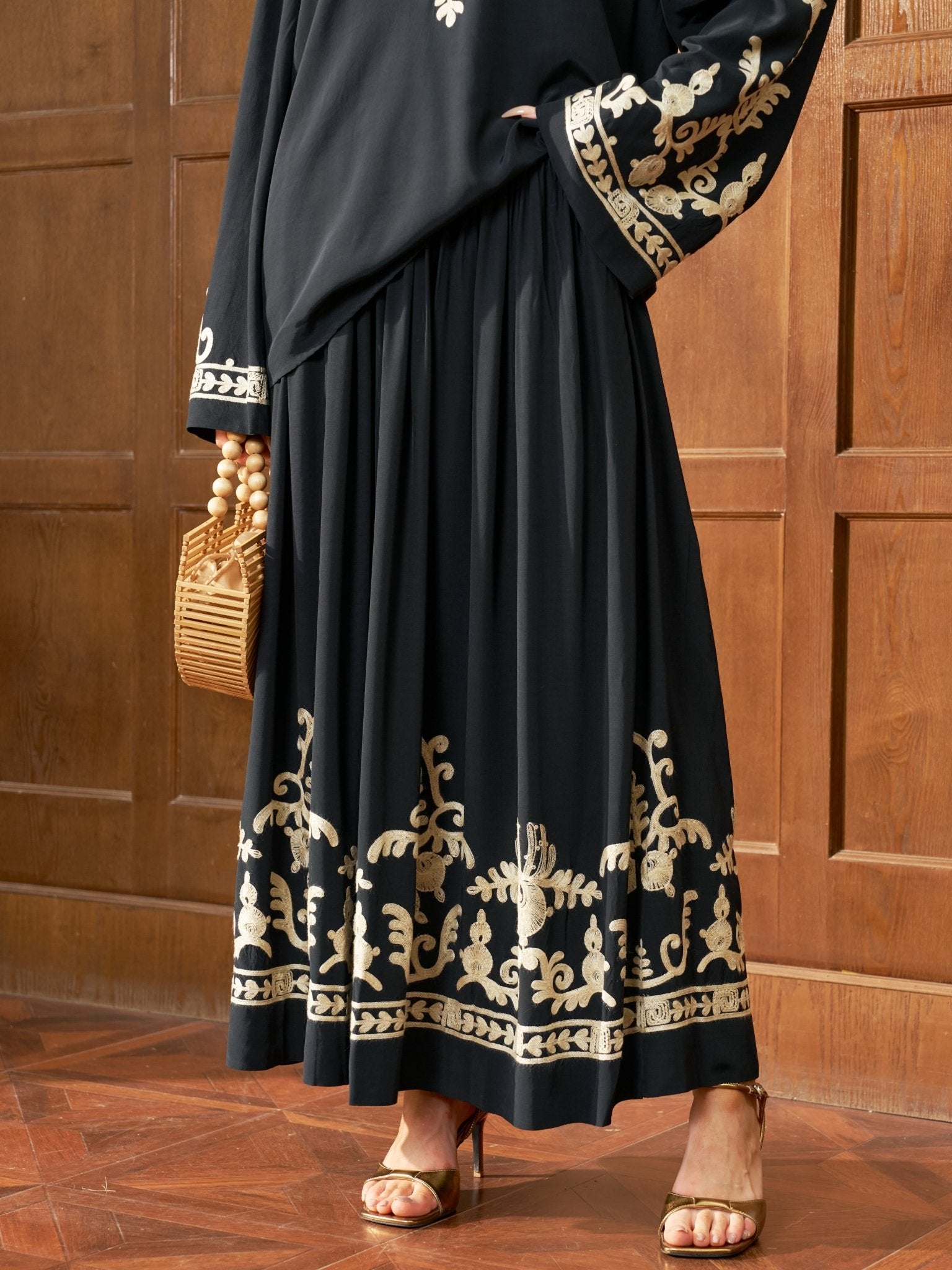 Wearing the Social Queen Contrast Embroidered A-Line Skirt, a person stands on a wooden floor by a paneled wall, holding a wicker handbag and wearing gold heeled sandals.