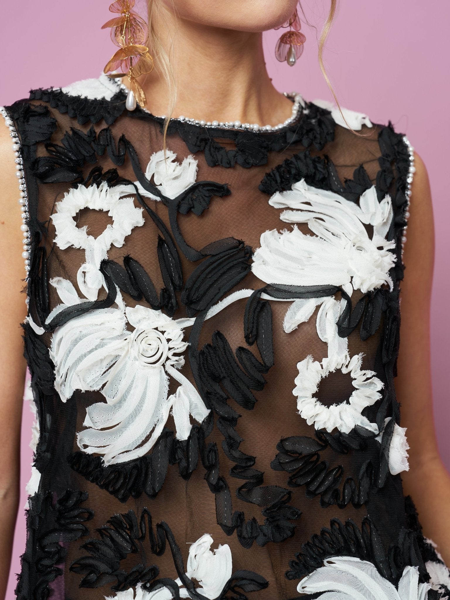 A model wears the Social Queen Contrast Floral Sheer Top, a sleeveless black mesh with bold white and black floral embroidery, set against a pink background.