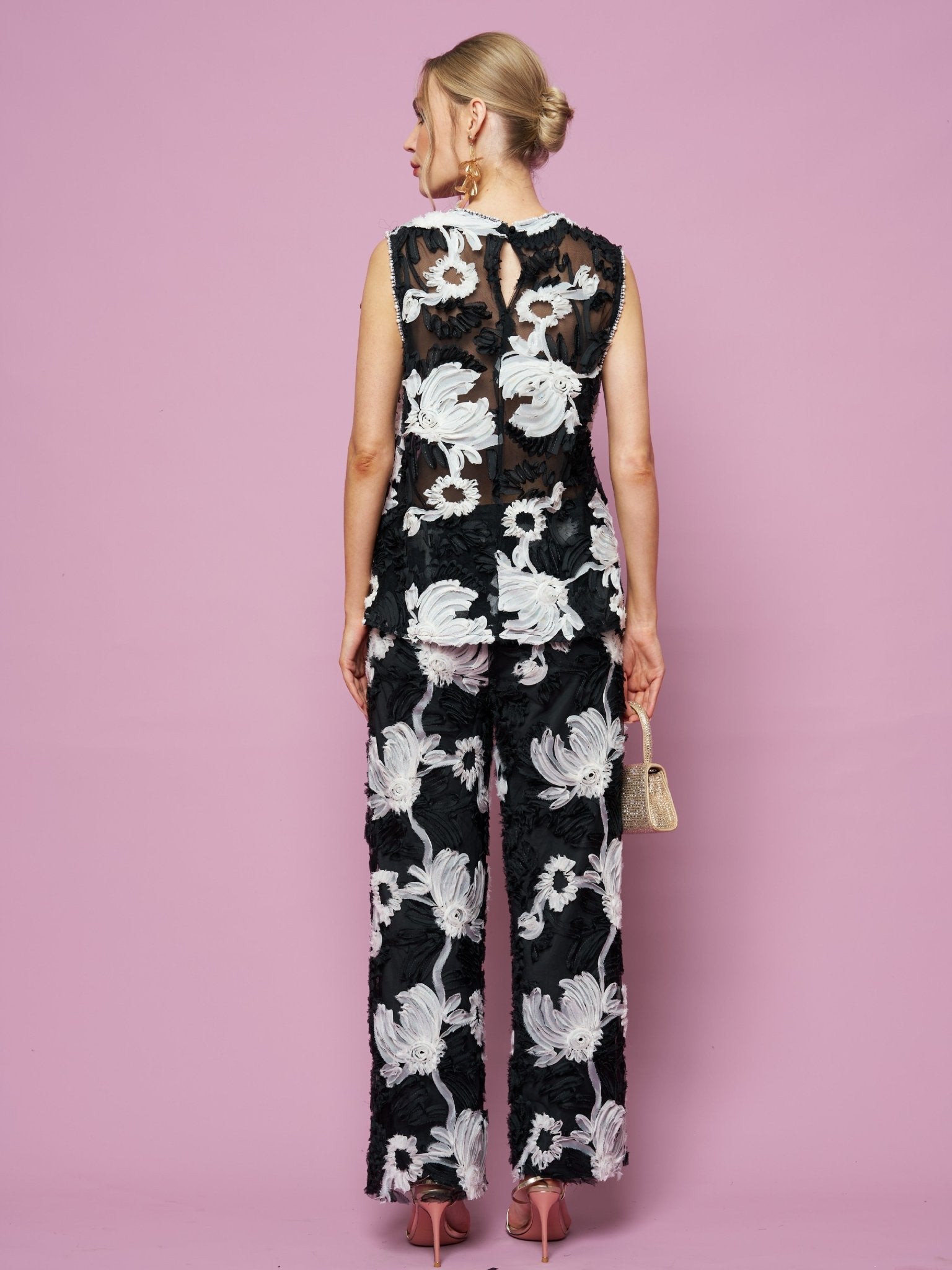 A woman stands facing away against a pink background, wearing Social Queen's Contrast Floral Sheer Wide-Leg Pants—a sleeveless black outfit with bold white floral patterns—while holding a small handbag.