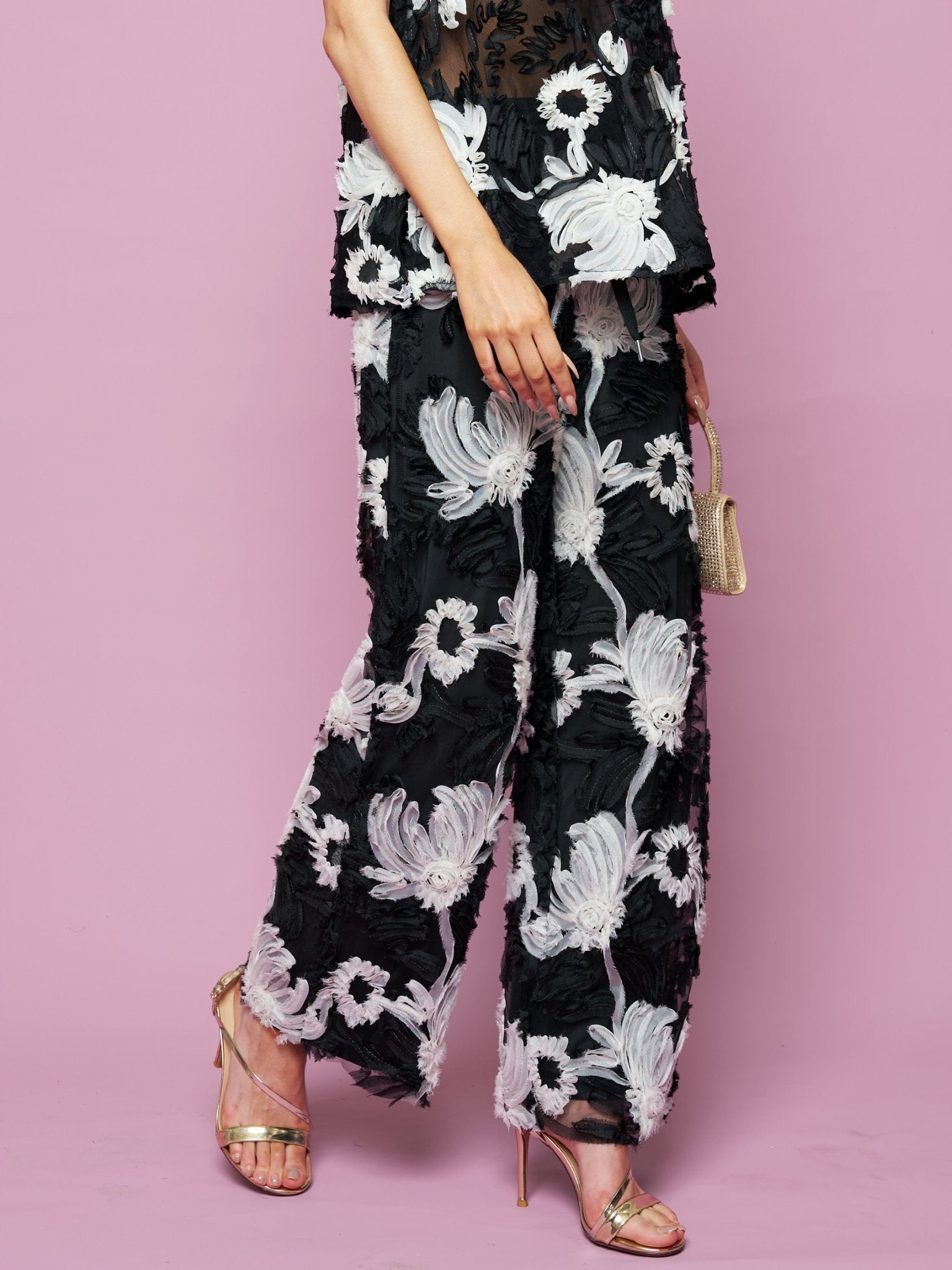 A person wears Social Queen’s Contrast Floral Sheer Wide-Leg Pants with a matching top, gold high heel sandals, and a small gold handbag, standing against a pink background.
