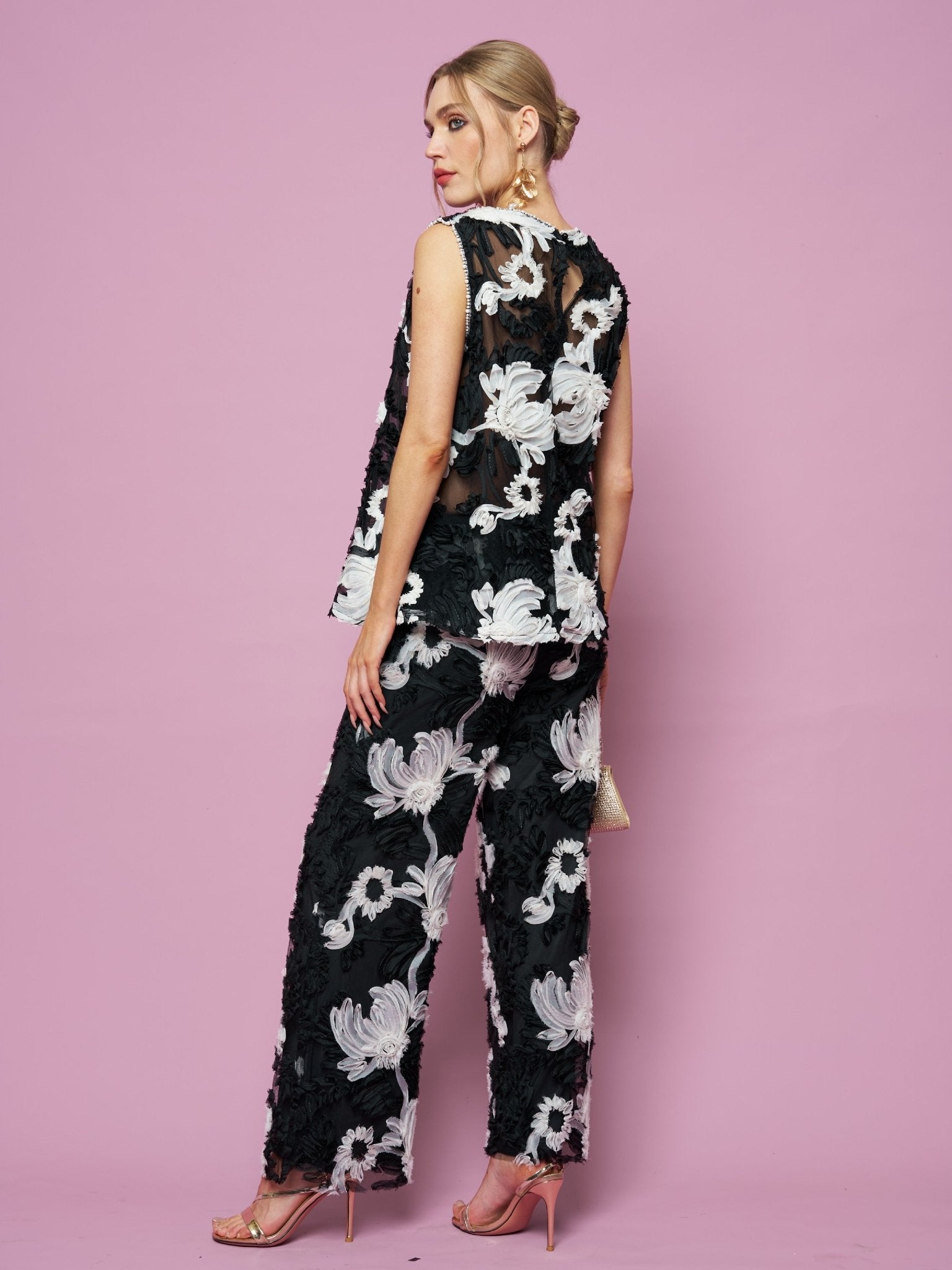 A woman poses against a pink backdrop in Social Queen's Contrast Floral Sheer Wide-Leg Pants, styled with a sleeveless black top, nude heels, and gold earrings.