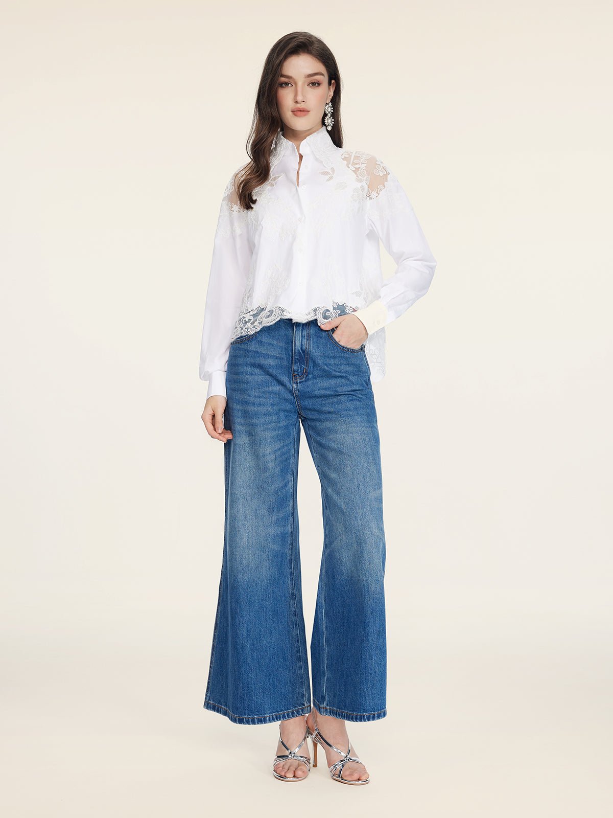 A woman faces forward in the Social Queen Cropped Embroidered Lace Panel Blouse, blue wide-leg jeans, silver heels, and drop earrings against a plain light background.