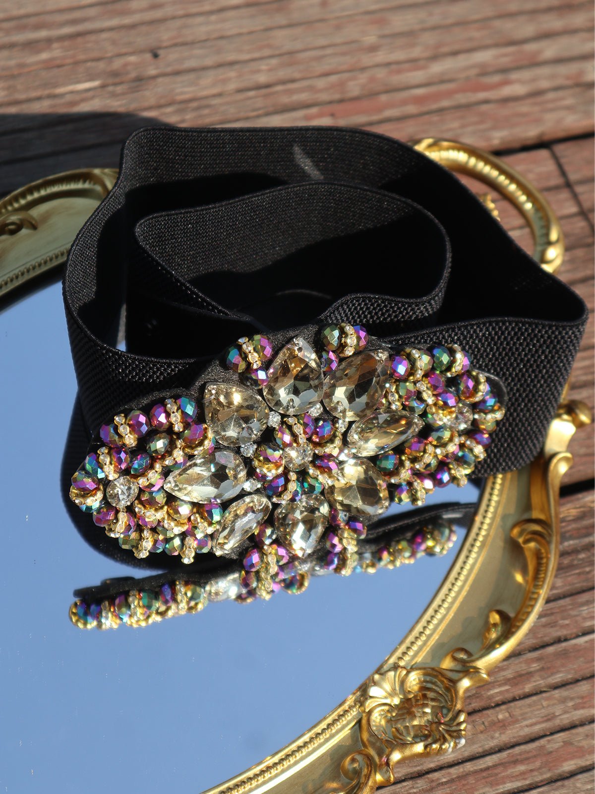 The Social Queen Crystal-Embellished Elastic Waist Belt with a jeweled multicolored buckle is showcased on an ornate gold mirror atop a wooden surface—a striking accessory for wedding guests.