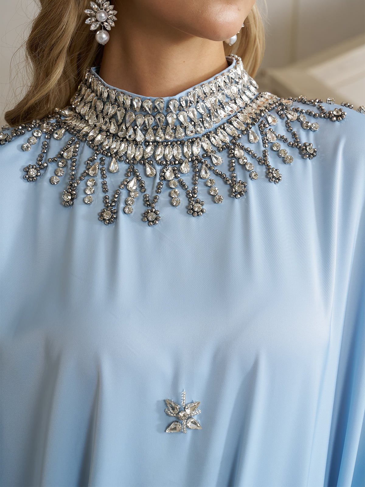 A woman wears the Social Queen Crystal-Embellished Mock-Neck Caftan Dress, featuring light blue fabric, an ornate jeweled collar, and a decorative chest brooch.