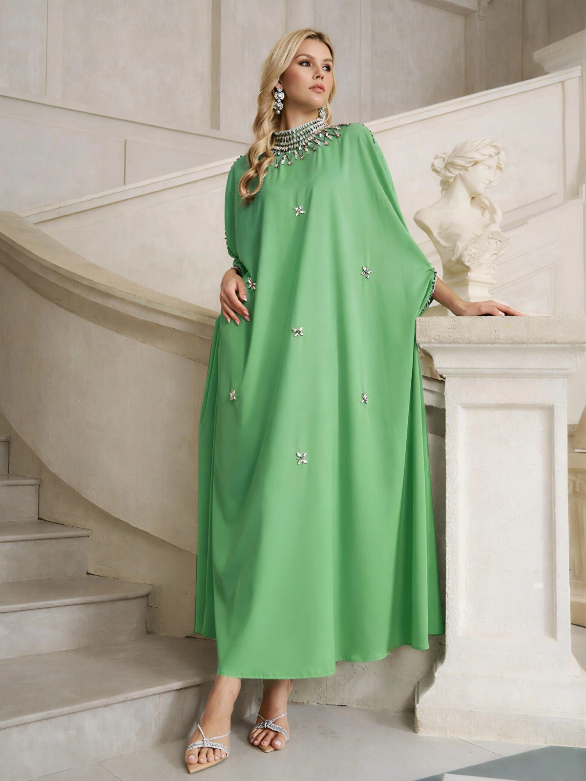 A woman wearing the Social Queen Crystal-Embellished Mock-Neck Caftan Dress stands on a staircase beside a white classical bust, one hand resting on the banister.