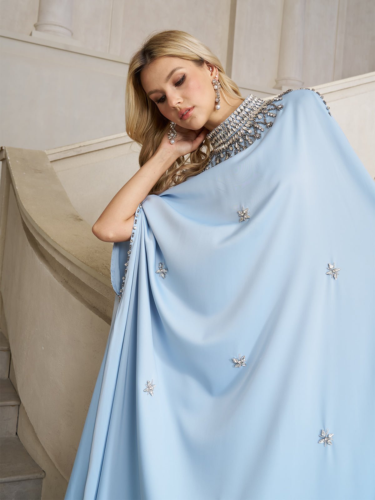 A woman in a Social Queen Crystal-Embellished Mock-Neck Caftan Dress stands on a staircase, resting her head on her hand and looking down.