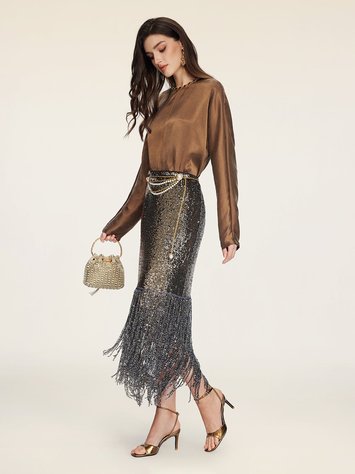 A woman stands against a plain background wearing the Social Queen Crystal-Embellished Tencel Satin Blouse, a metallic fringed skirt, gold heels, and holding a beaded purse.