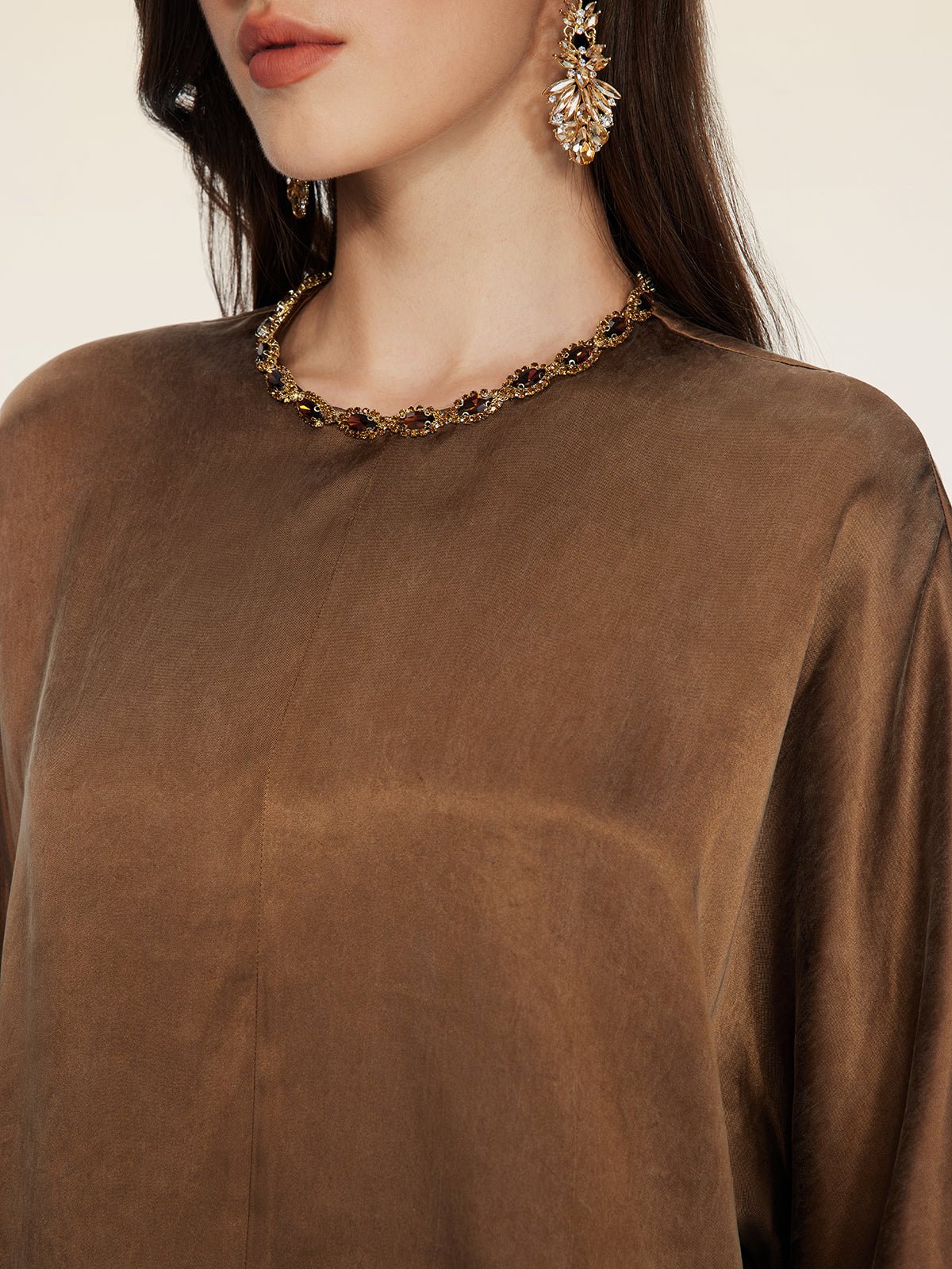A woman shown from the shoulders up wears Social Queen's Crystal-Embellished Tencel Satin Blouse, a necklace with hand-set crystals, and a large decorative earring.