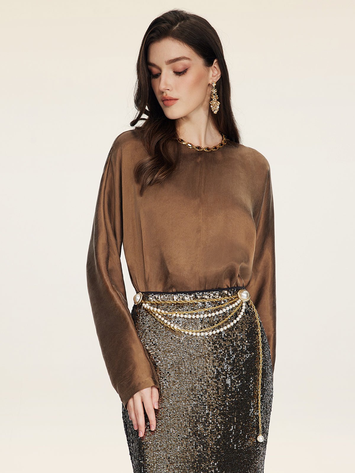 Woman wearing the Social Queen Crystal-Embellished Tencel Satin Blouse, styled with a gold chain necklace, high-waisted black sequin skirt, pearl and chain belt, and gold drop earrings.