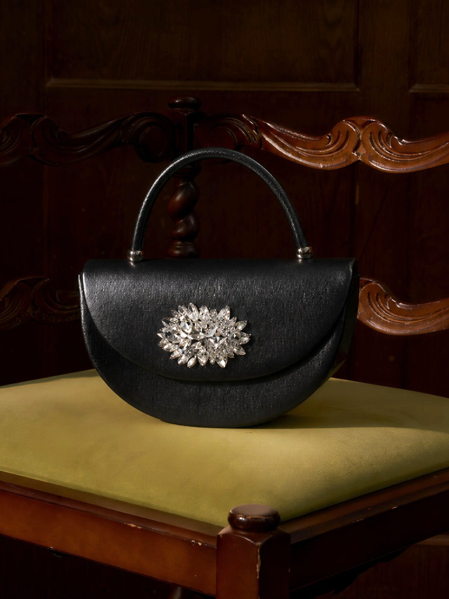 The Social Queen Crystal Floral Satin Mini Top-Handle Bag with a jeweled brooch sits on a yellow-cushioned, carved wooden chair.