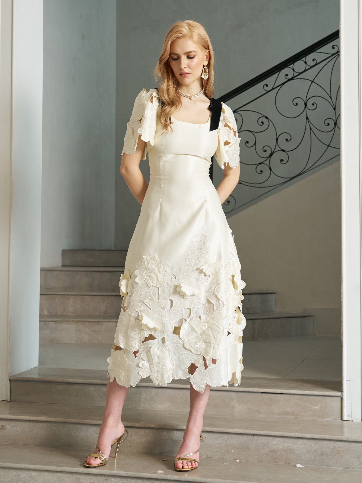 A woman in the Social Queen Cutout Floral Appliqué Satin Dress with a petal hem stands on an indoor staircase, looking down with her hands in front of her.