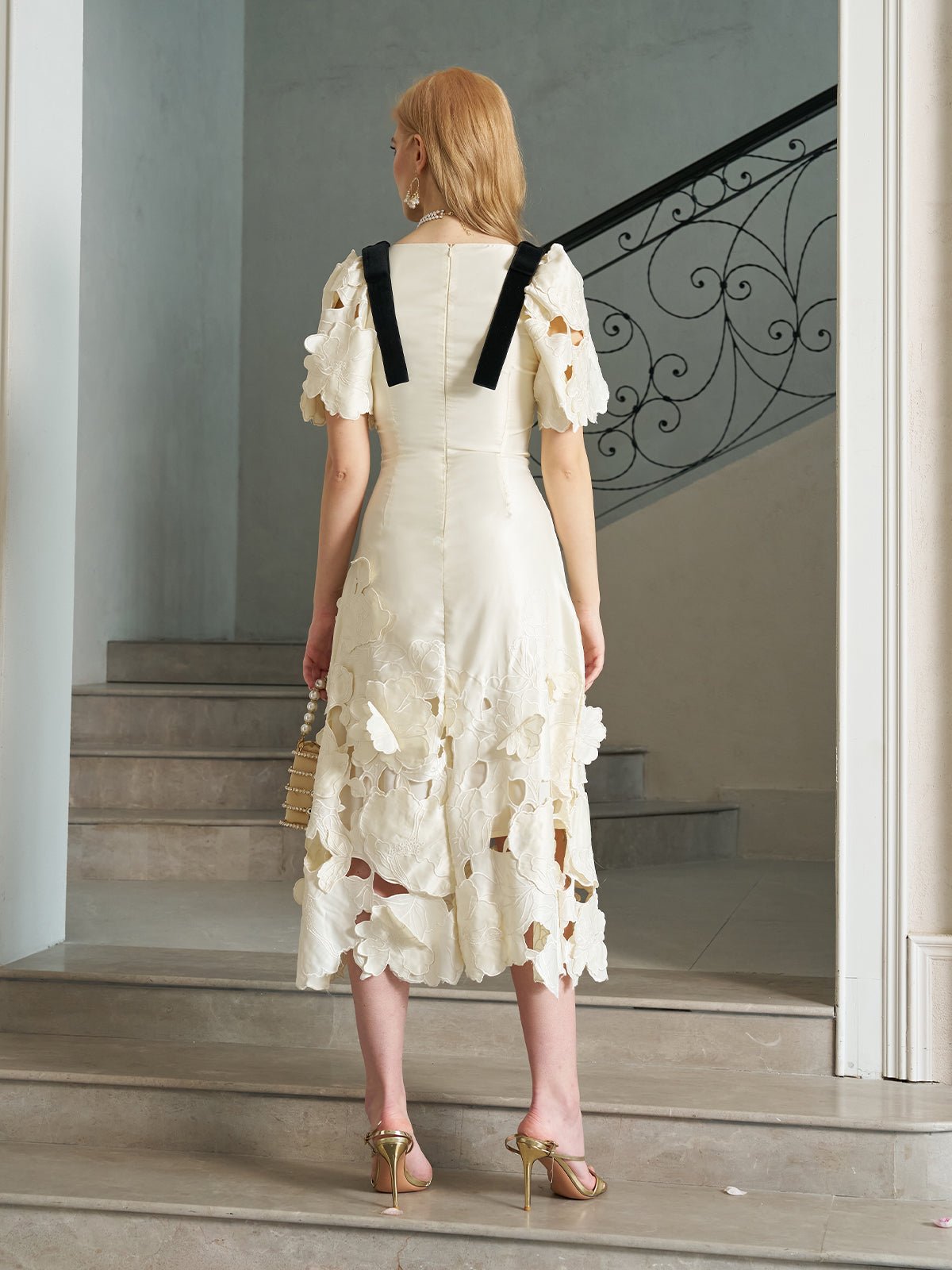 A woman stands on a staircase facing away, wearing the Social Queen Cutout Floral Appliqué Satin Dress with black straps and petal hem, paired with gold heels and holding a small woven bag.