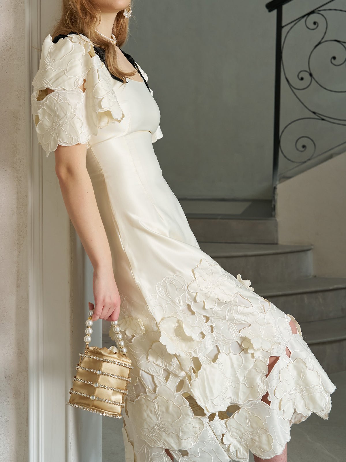A woman in the Social Queen Cutout Floral Appliqué Satin Dress holds a small, pearl-handled handbag while standing by a staircase.