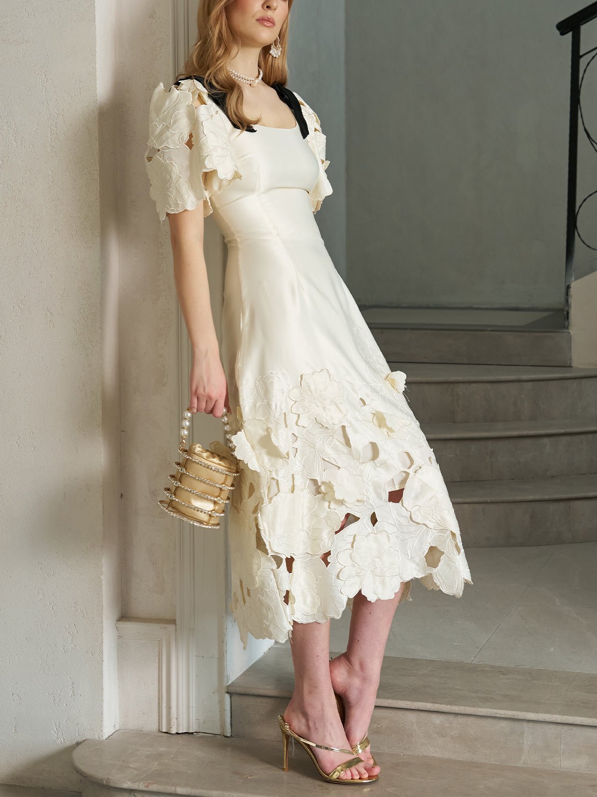 A woman in the Social Queen Cutout Floral Appliqué Satin Dress in ivory stands on a staircase, holding a gold handbag and wearing gold high-heeled sandals.