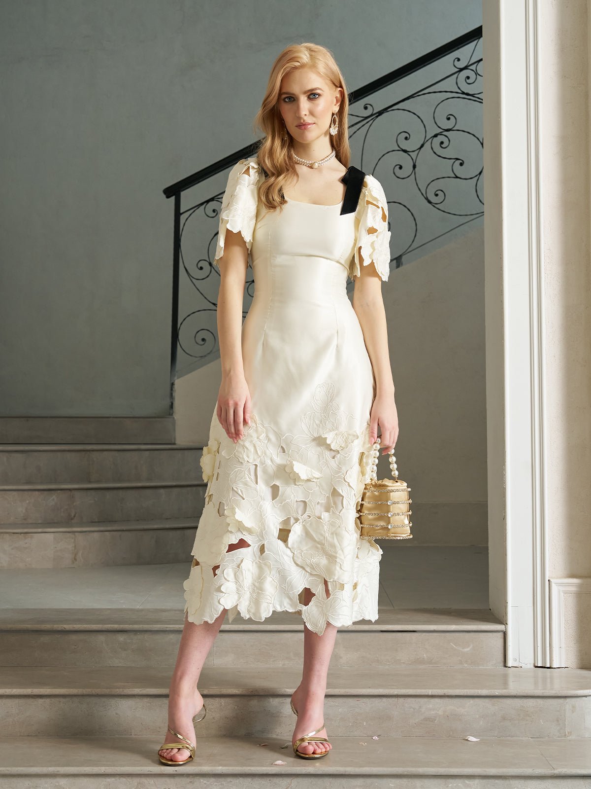 A woman stands on stone steps with a wrought iron railing, wearing the Social Queen Cutout Floral Appliqué Satin Dress in ivory, paired with strappy sandals and a small handbag.
