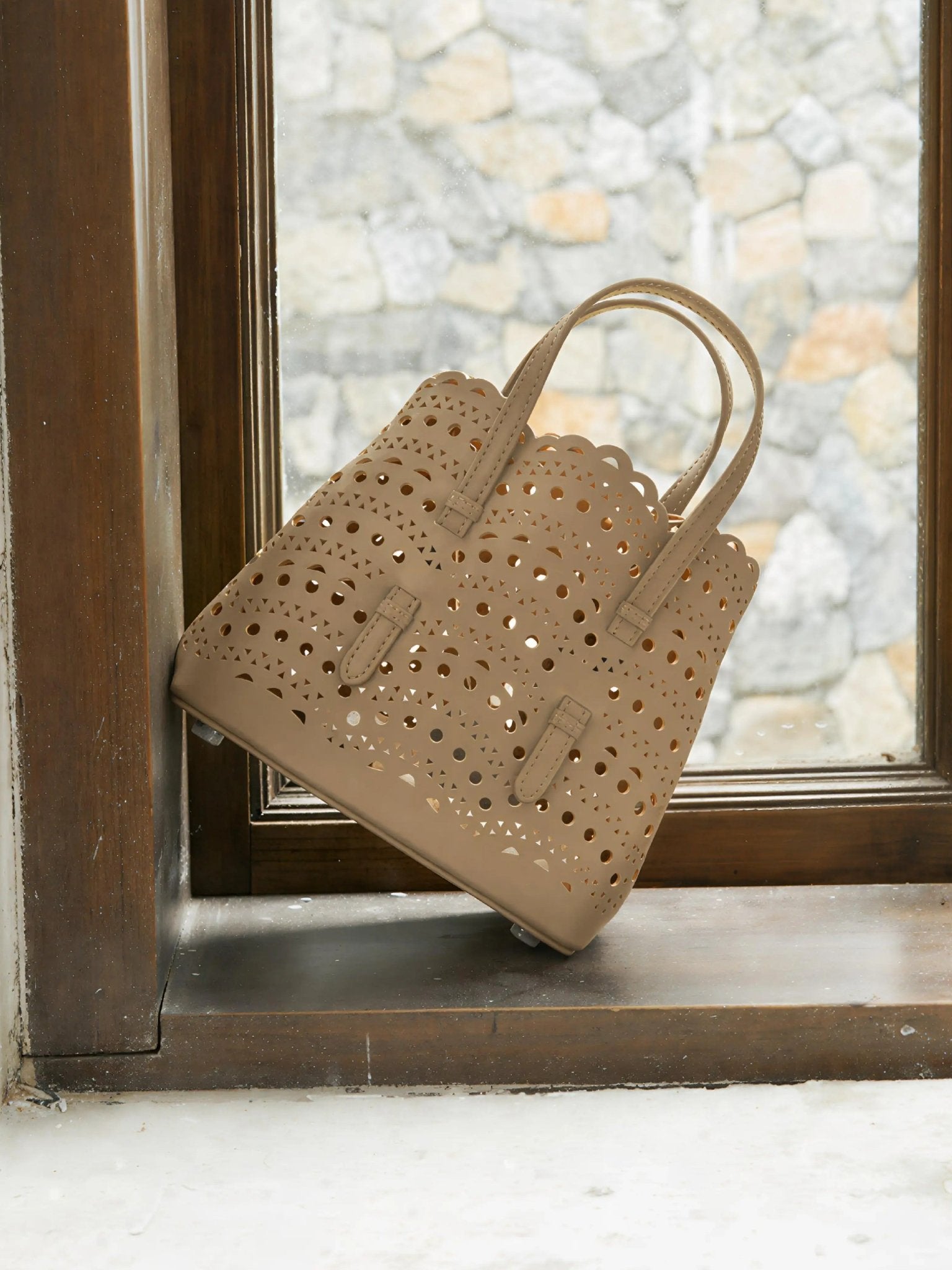 The Social Queen Cutout Leather Top-Handle Bag, featuring a tan geometric cutout design and double handles, rests on a wooden window ledge with a stone wall visible outside.