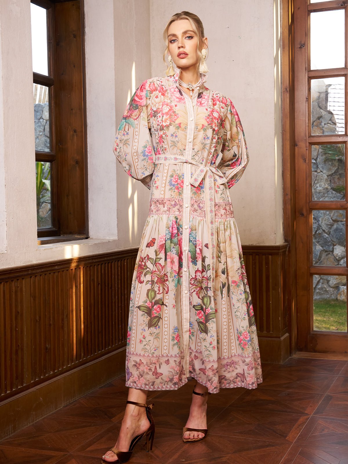 A woman stands indoors by wooden windows, wearing the Eden Garden Print Tie-Waist Dress by Social Queen with sheer sleeves and dark heels—an elegant choice for a wedding guest dress.