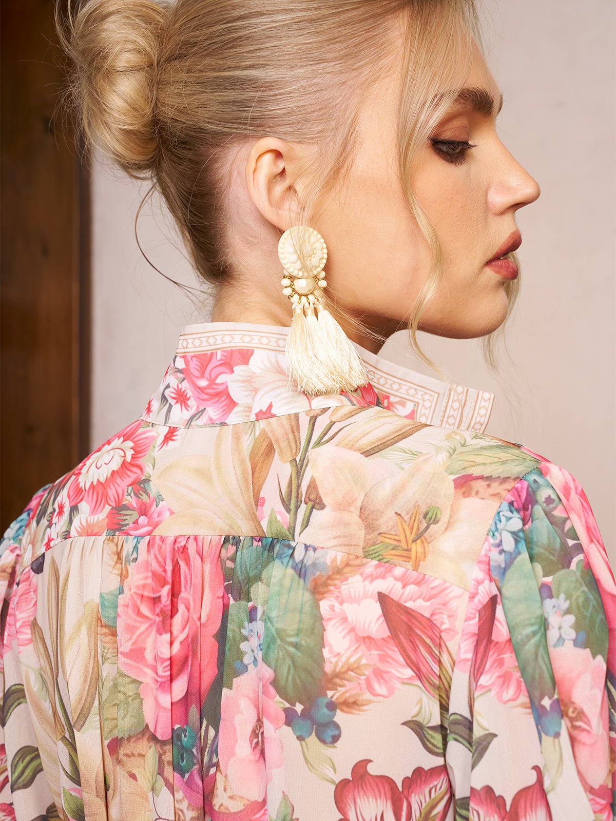 Woman with blonde hair in a bun wearing large tassel earrings and the Eden Garden Print Tie-Waist Dress by Social Queen, featuring a sheer silk-look pastel floral pattern, styled elegantly as a wedding guest dress, shown from side and back.