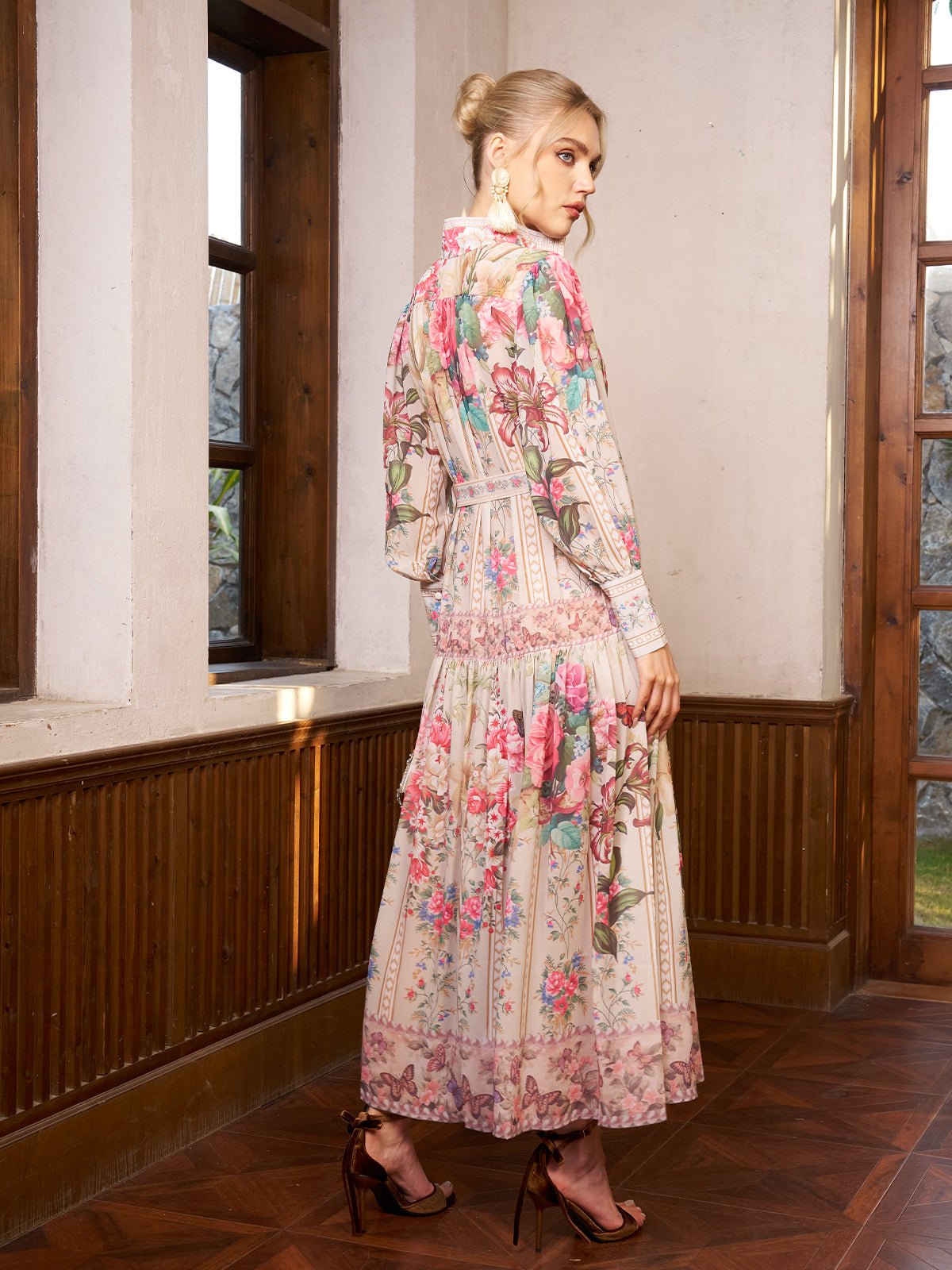A woman wearing the Eden Garden Print Tie-Waist Dress by Social Queen stands indoors in high heels, facing away from the camera near tall wooden-framed windows. The dress features long sleeves and a garden print.