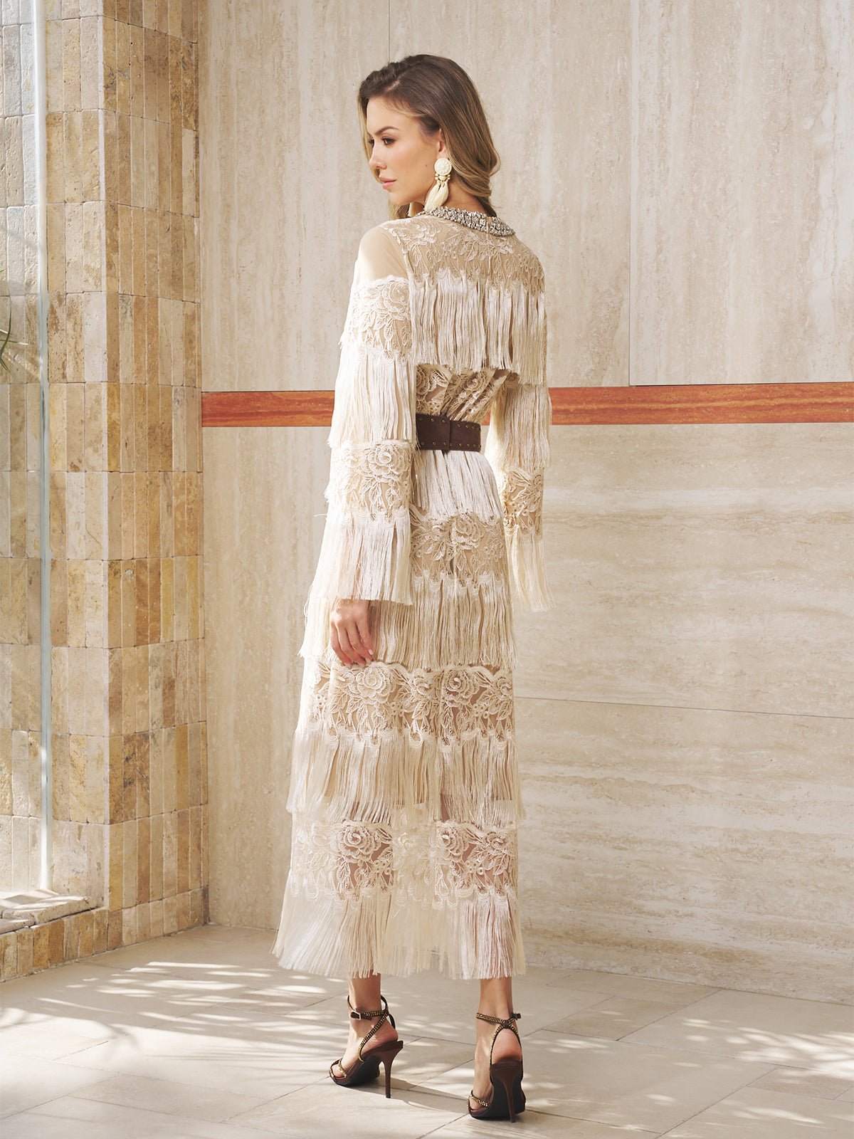 A woman stands indoors in a Social Queen Embroidered Fringe Lace Crystal-Trimmed Dress, featuring a long, sheer cream design with lace, fringe details, and floral embroidery. She pairs it with high heels and statement earrings.