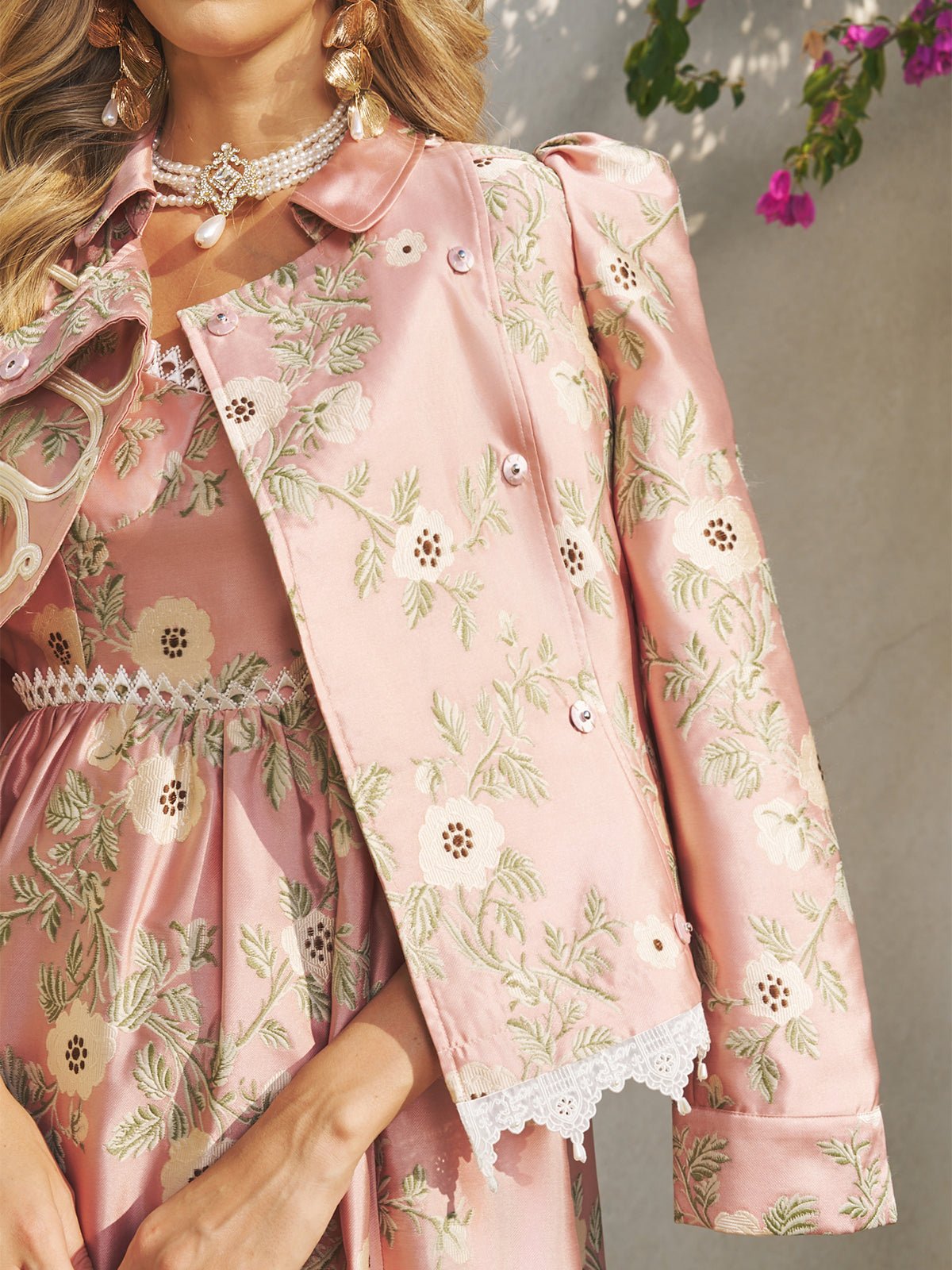 A woman wears the Social Queen Embroidered Jacquard Lace-Trimmed Jacket over a pink floral dress, accessorized with pearl jewelry—an elegant wedding guest look.