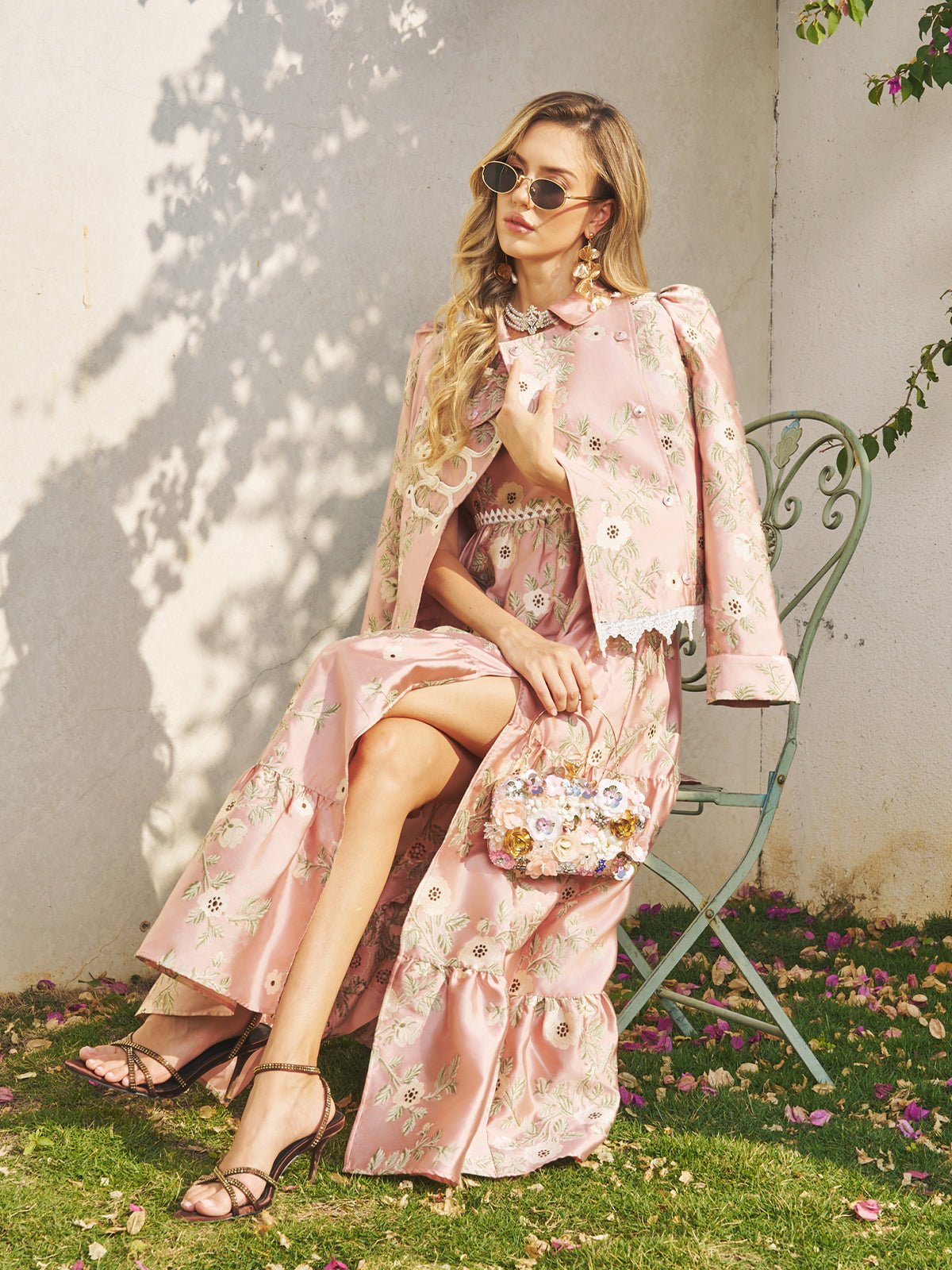 Woman in a pink floral dress and Social Queen Embroidered Jacquard Lace-Trimmed Jacket sits outdoors on a green metal chair, accessorized with sunglasses and a small decorative purse—an elegant choice for a wedding guest.