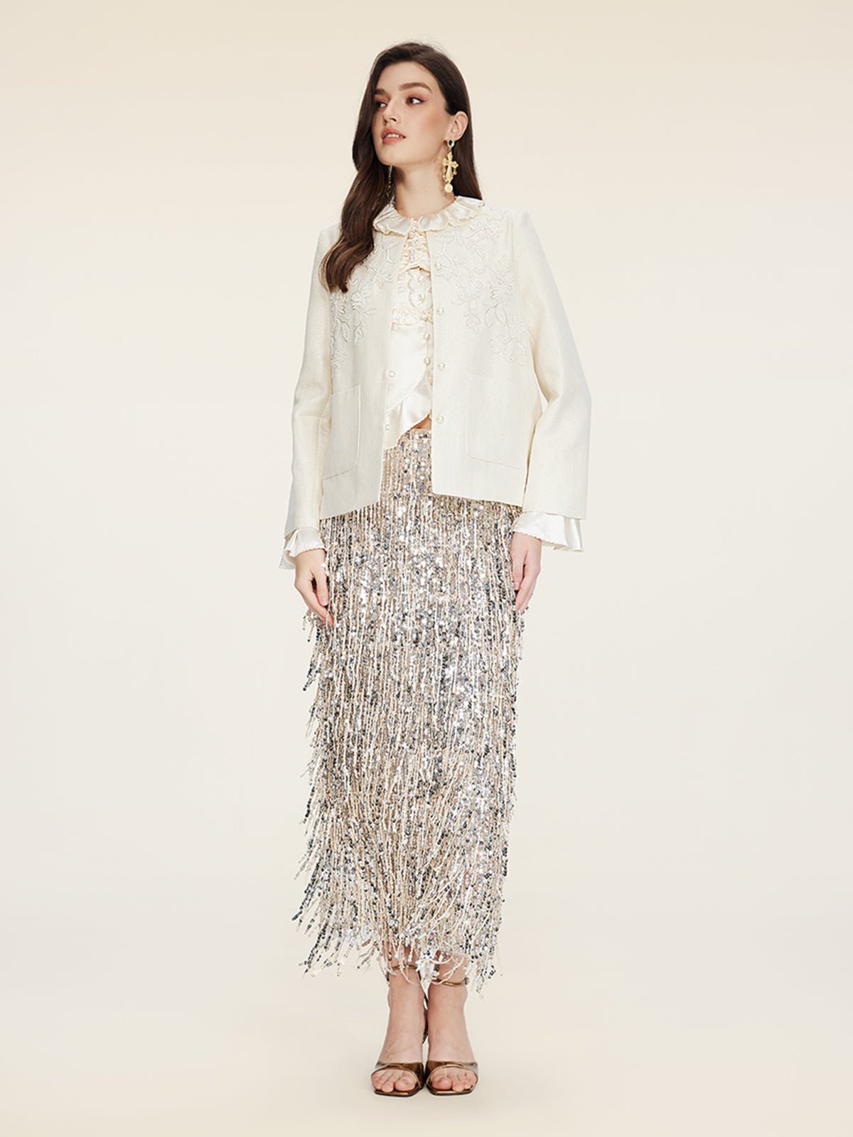 A woman stands against a plain background wearing Social Queen’s Embroidered Wool Jacket with Pearl Accents, a blouse, and a silver fringed midi skirt with high-heeled sandals.