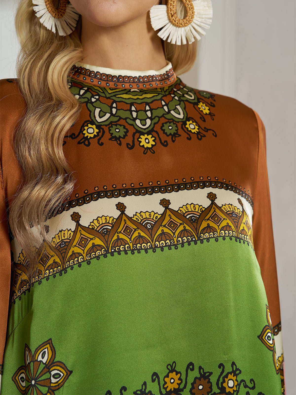 A woman wears the Social Queen Exotic Placed Print Silk Slit Dress, featuring long sleeves and brown-green mandala patterns, styled as chic resort wear with large circular earrings accented by white fringe.