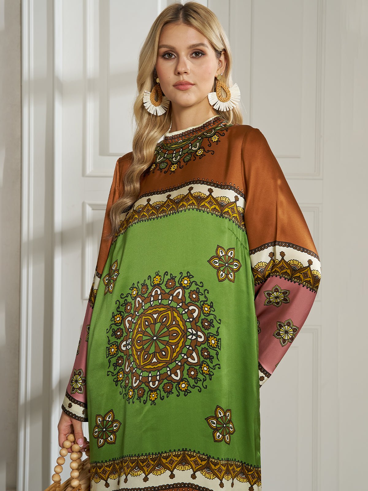 A woman indoors wears the Social Queen Exotic Placed Print Silk Slit Dress, featuring green, brown, and pink mandala floral patterns. Ideal for resort wear, she pairs it with a beaded bag and statement earrings.