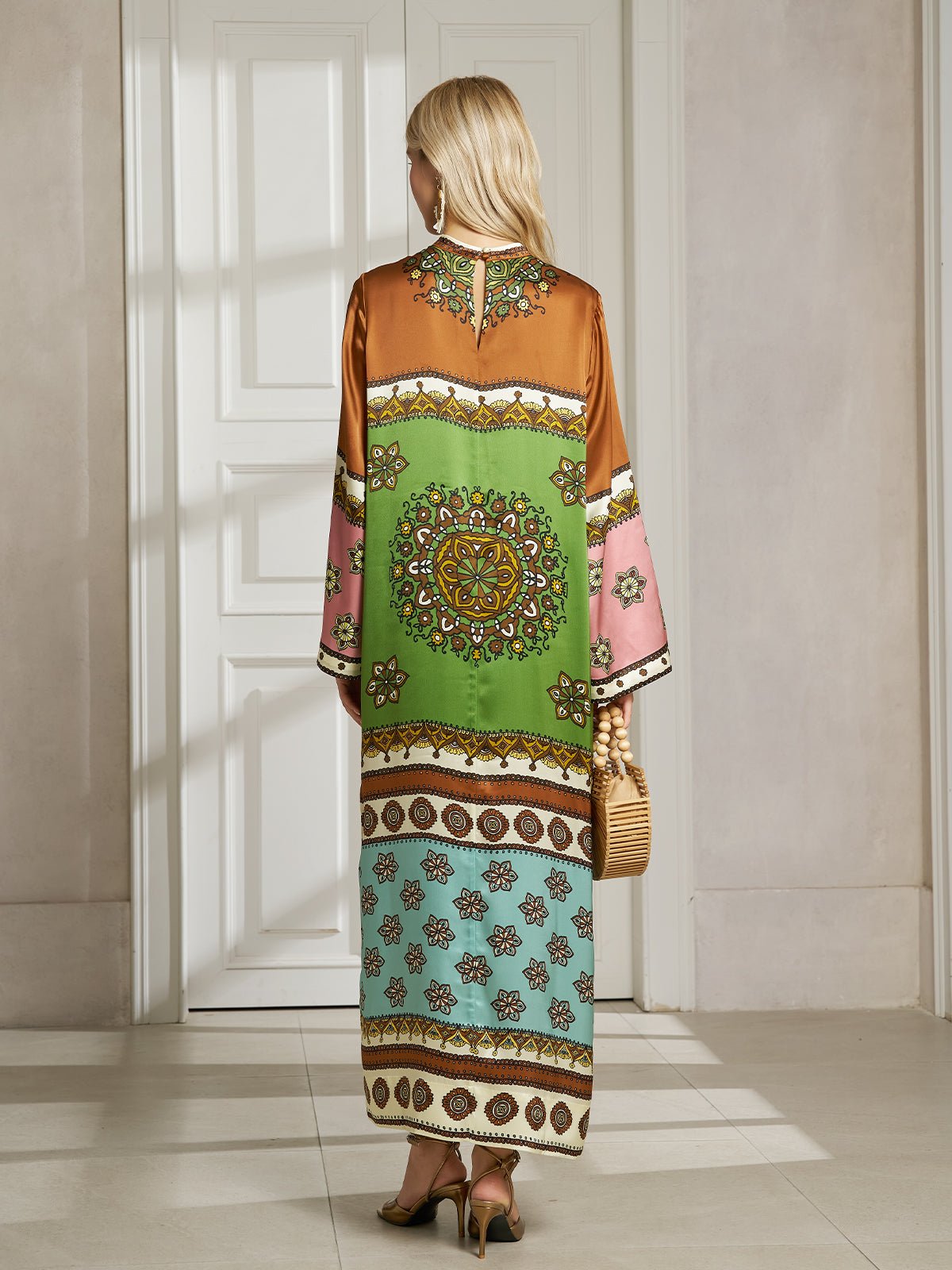 A woman in heels faces a white door, wearing the Social Queen Exotic Placed Print Silk Slit Dress with green, brown, pink, and blue mandala-inspired floral sections and carrying a small woven handbag—perfect resort elegance.