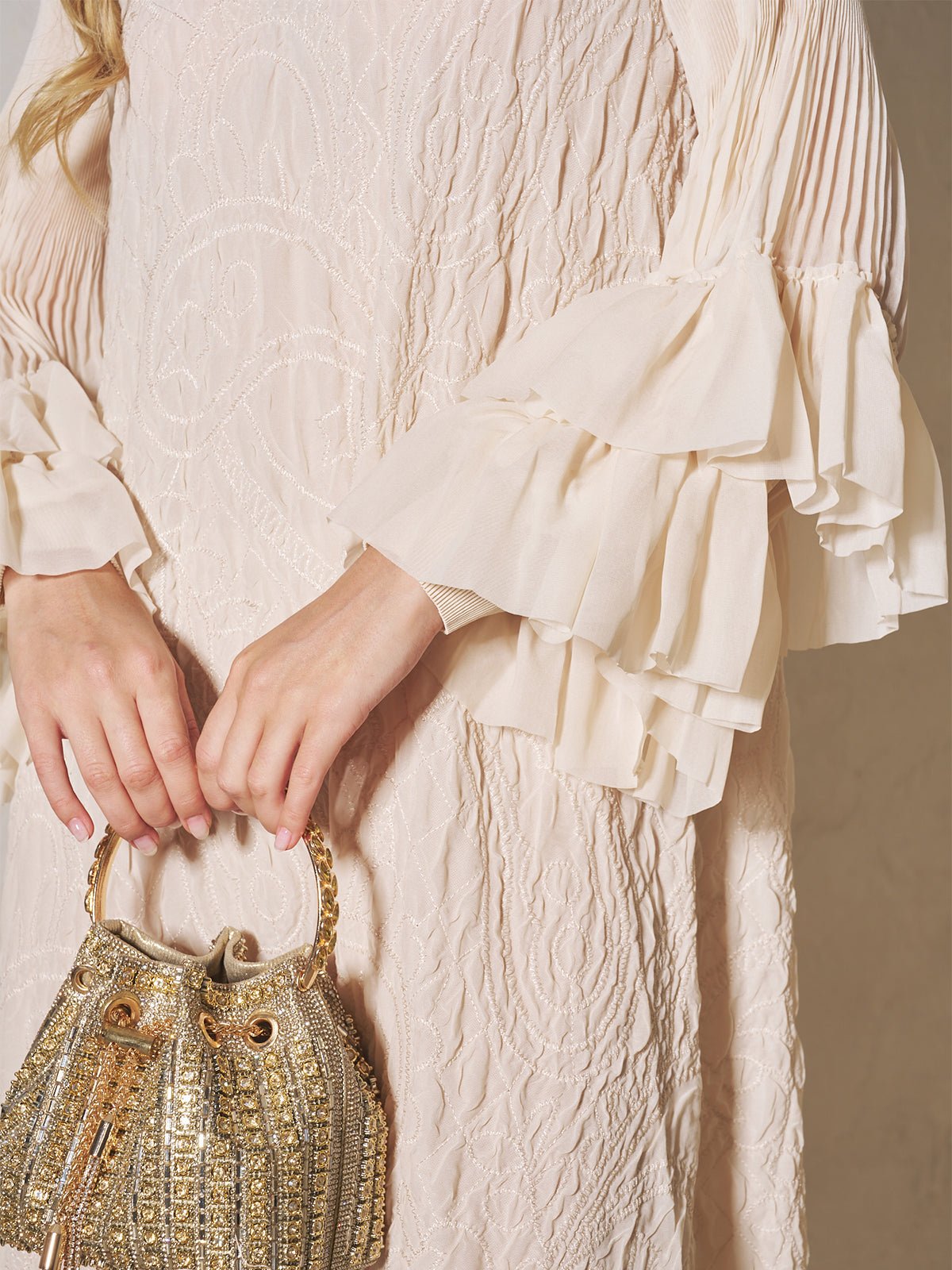 A woman wears the Social Queen Floral Appliqué Embroidered Ruffle Dress, featuring textured cream lace and ruffled sleeves, while holding a gold beaded handbag with both hands.