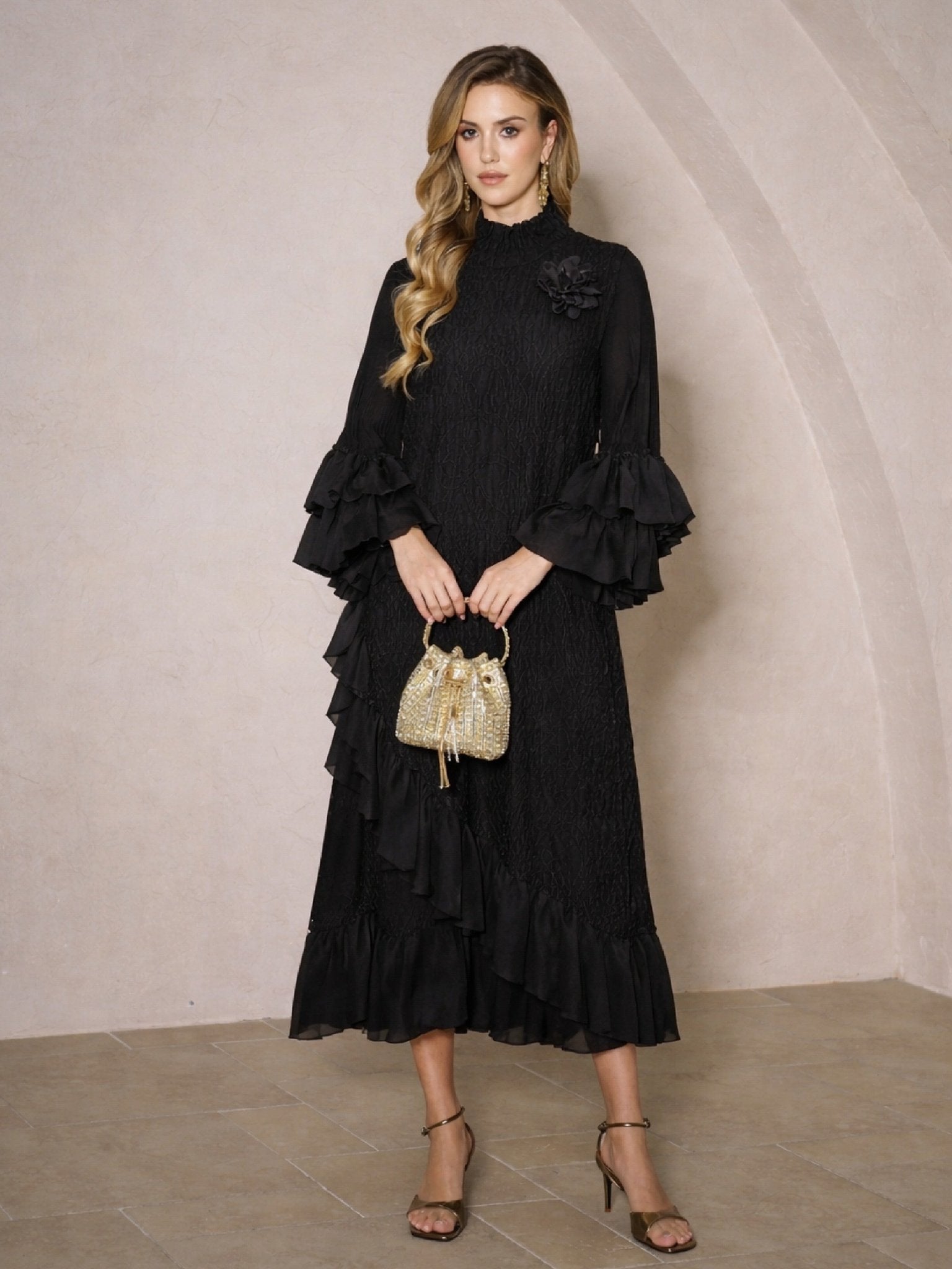 A woman stands indoors in a Social Queen Floral Appliqué Embroidered Ruffle Dress, holding a small gold handbag and wearing high-heeled sandals.