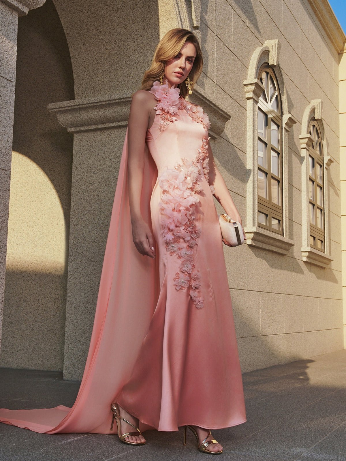 A woman stands outside a stone building holding a clutch, wearing the Social Queen Floral Appliqué Satin Fishtail Gown—a sleeveless pink satin dress with 3D floral details and a cape.
