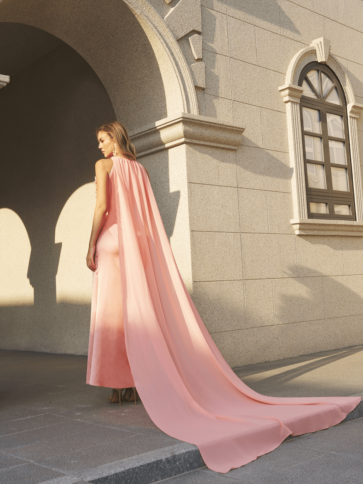 A woman stands outside a stone building in the Social Queen Floral Appliqué Satin Fishtail Gown, featuring a peach hue, 3D floral appliqués, and a floor-length cape.