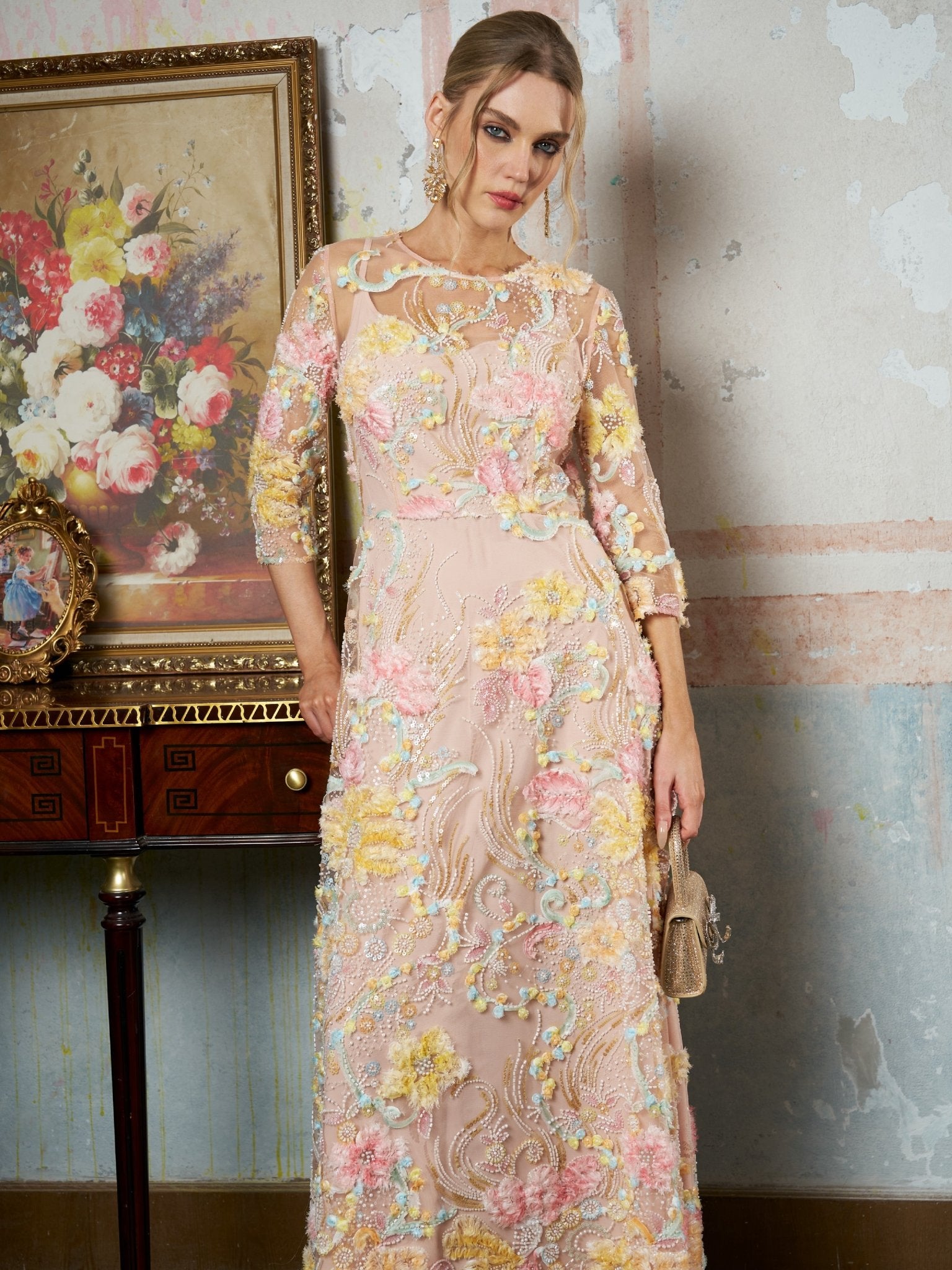 A woman indoors stands by a table with framed art and flowers, holding a small gold handbag. She is wearing the Social Queen Floral Beaded Sheer Gown—a long, pastel A-line evening dress with 3D floral appliqué.