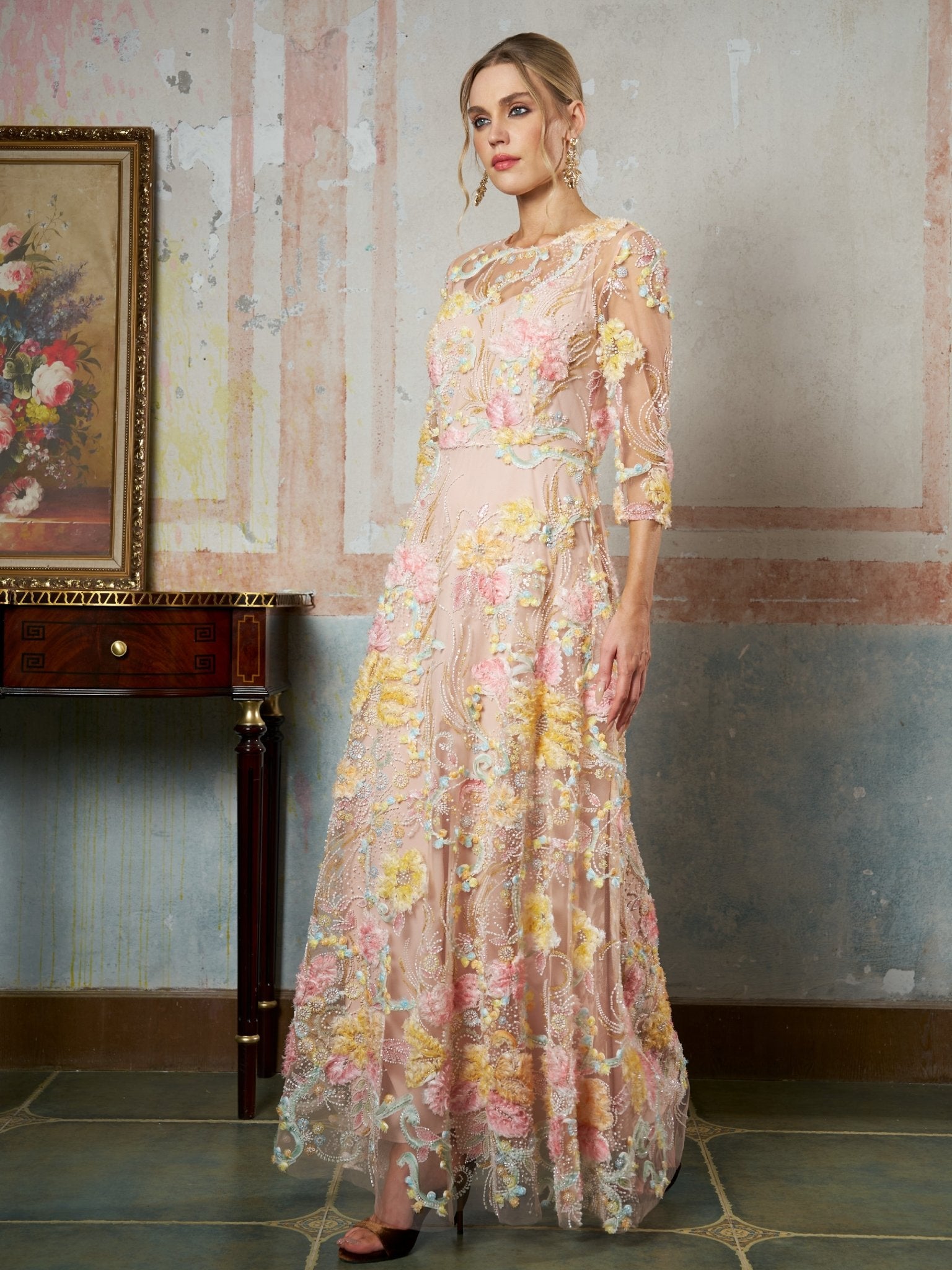 A woman stands indoors in the Social Queen Floral Beaded Sheer Gown, featuring an A-line silhouette, floral embroidery, and three-quarter sleeves, beside a wooden table and a floral painting.