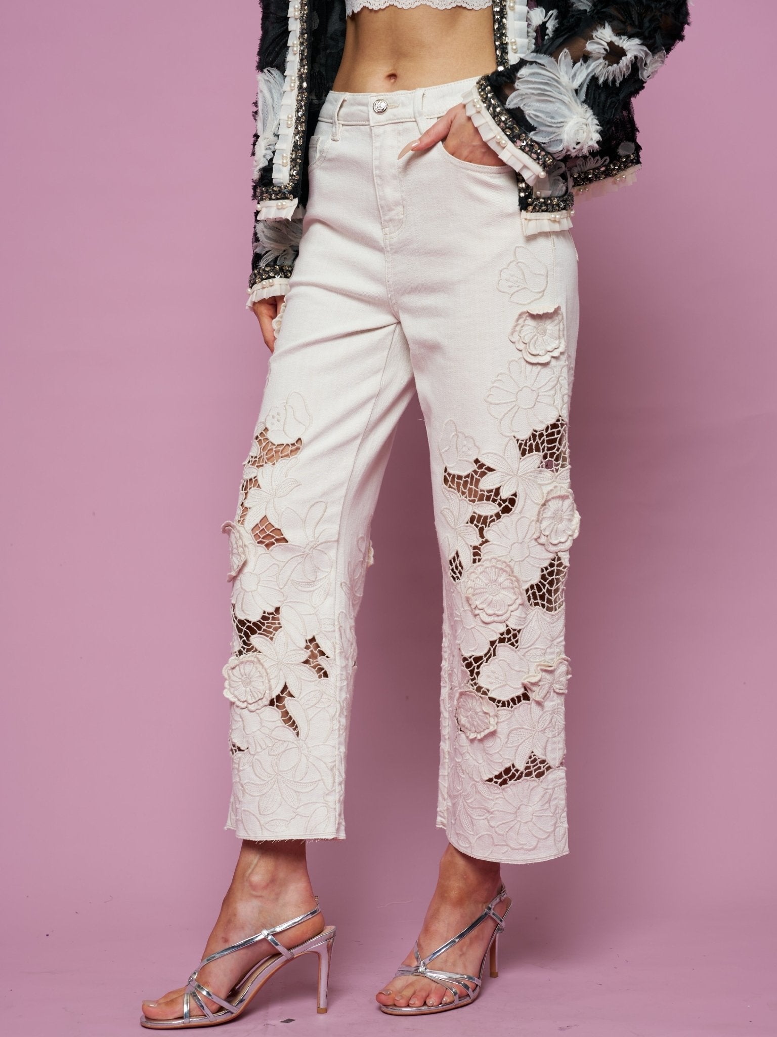 Wearing Social Queen’s Floral Cutout High-Rise Jean, a crop top, black embellished jacket, and strappy high-heeled sandals, a person poses against a pink background.