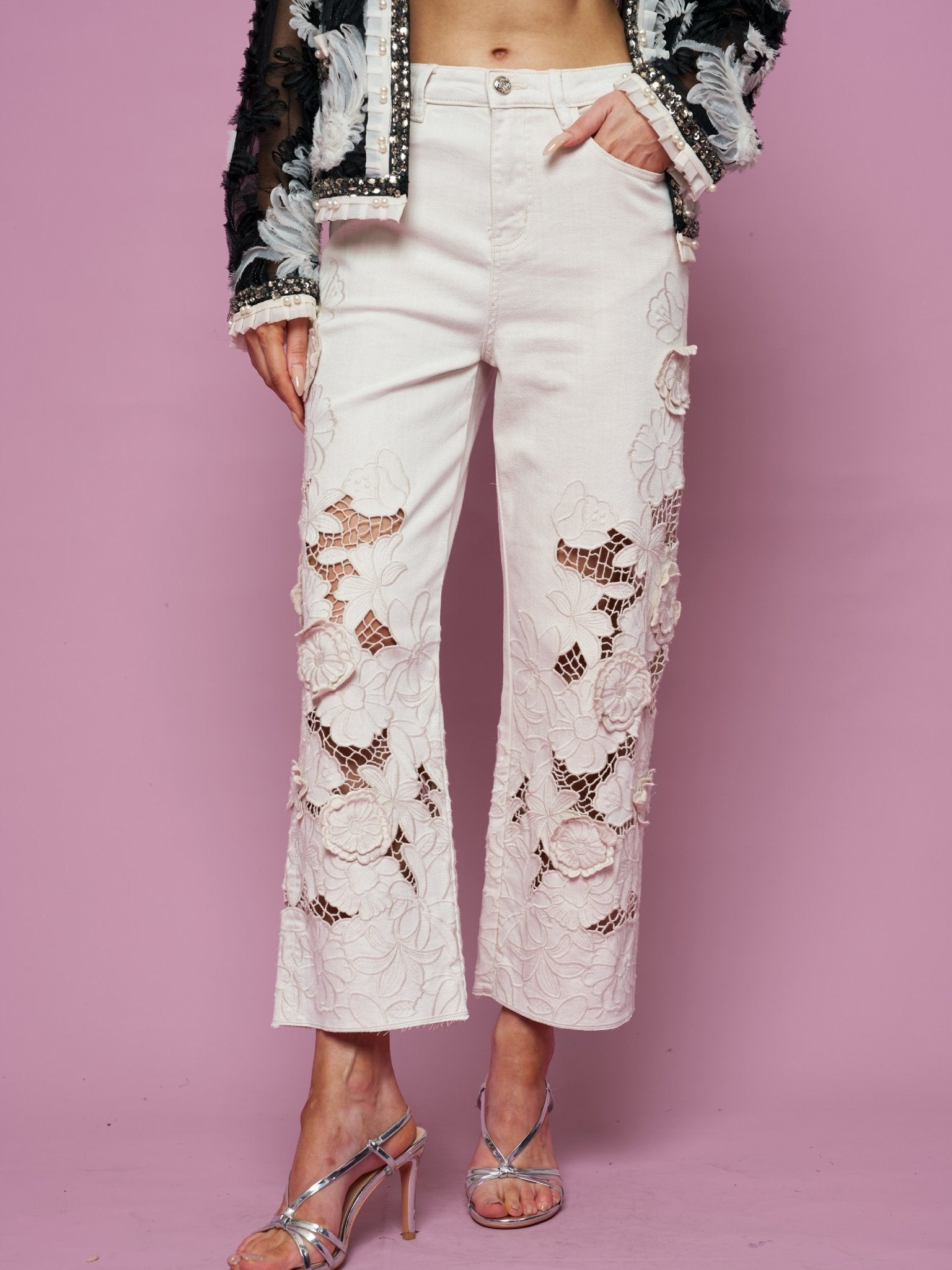 A person in Social Queen’s Floral Cutout High-Rise Jean, a black and white textured jacket, and silver high-heeled sandals stands against a pink backdrop.
