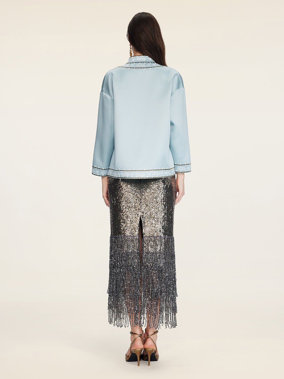 A woman stands with her back to the camera, wearing the Social Queen Floral Embroidered Beaded Jacket and a silver sequin fringe skirt, styled with gold heeled sandals.