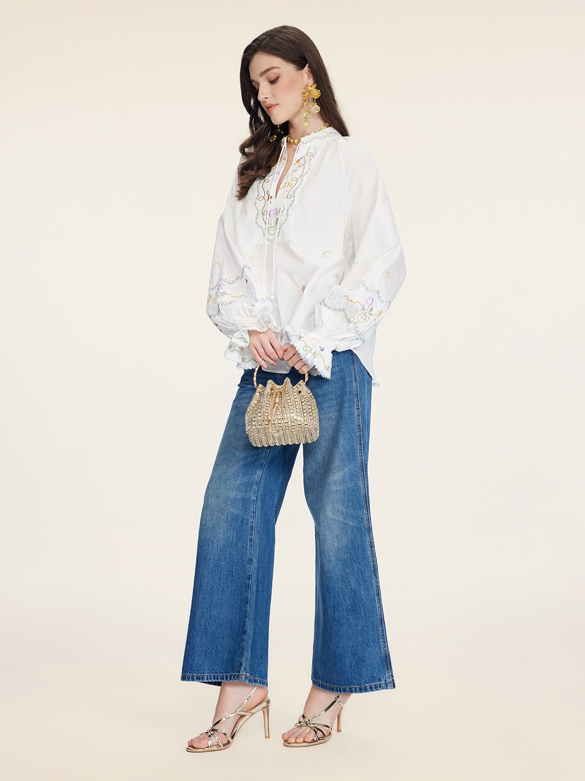A woman stands against a plain background wearing the Social Queen Floral Embroidered Cotton Blouse, blue wide-leg jeans, metallic heels, gold earrings, and holding a beaded handbag.