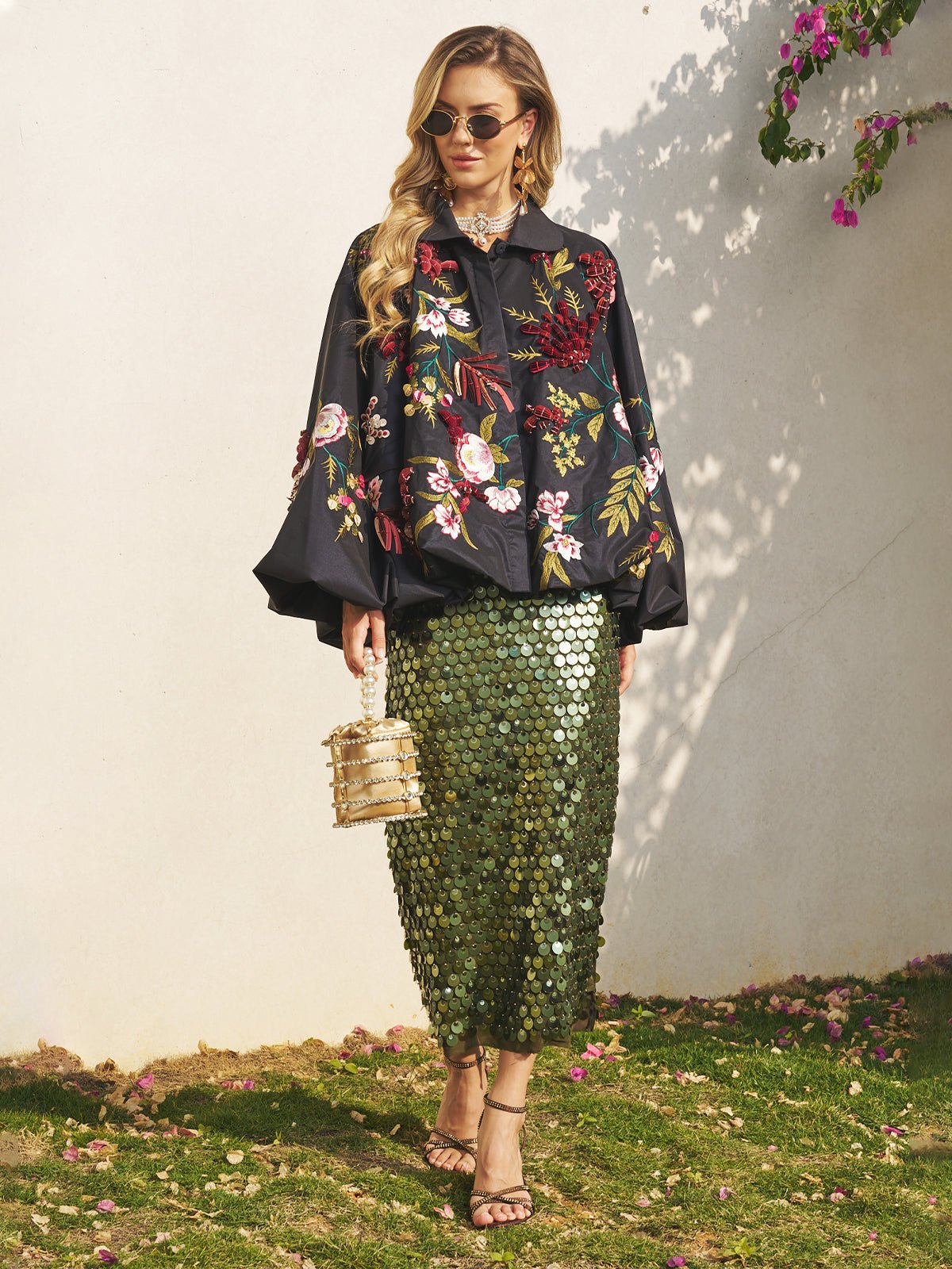 A woman in sunglasses, a Social Queen Floral Embroidered Floral Puff Sleeve Jacket, green sequin skirt, sandals, and basket bag—perfect for a wedding guest—stands on grass by a white wall with pink flowers overhead.